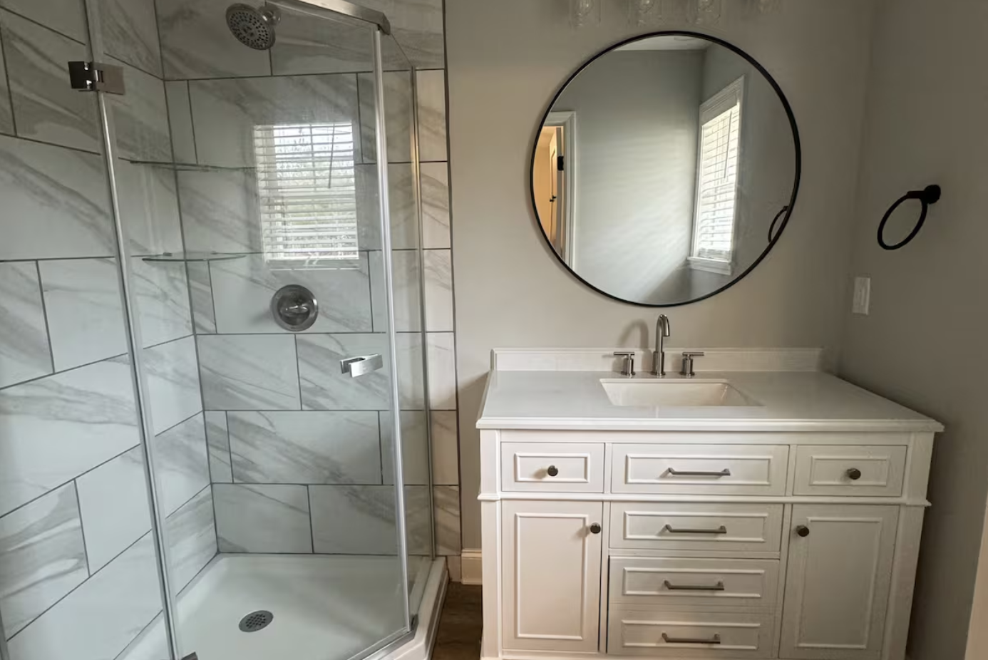 Bathroom with a glass shower enclosure on the left and a white vanity with a sink, faucet, and mirror on the right; a window with blinds is reflected in the mirror.