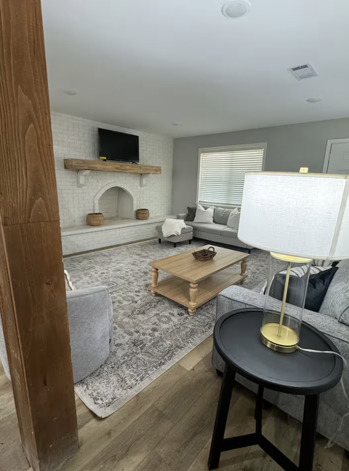 Living room with a white brick fireplace, a wooden mantle, and a TV above it. There is a sofa, an armchair, and a coffee table. A large window with blinds is on one wall, and a table lamp is on a side table. The room has wood flooring and a patterned