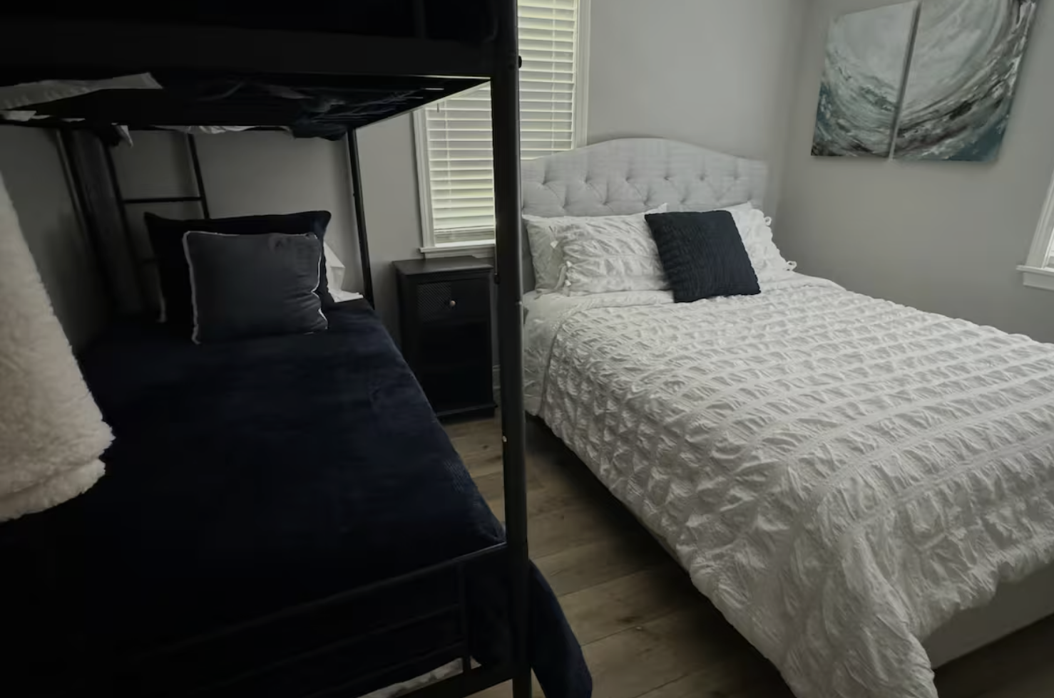 A bedroom with a white bed with a padded headboard, black and white pillows, a black nightstand, and a window with blinds. A bunk bed with black bedding and pillows is partially visible on the left side of the image. A set of three abstract art paint