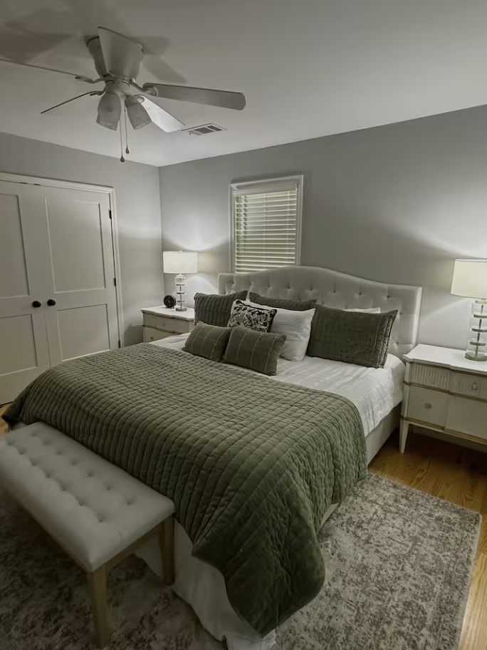 A cozy bedroom with a white tufted headboard, green quilted bedding, and multiple pillows. There are two matching white nightstands with lamps, a window with blinds, a ceiling fan, and a white door. A small bench sits at the foot of the bed, and a ru