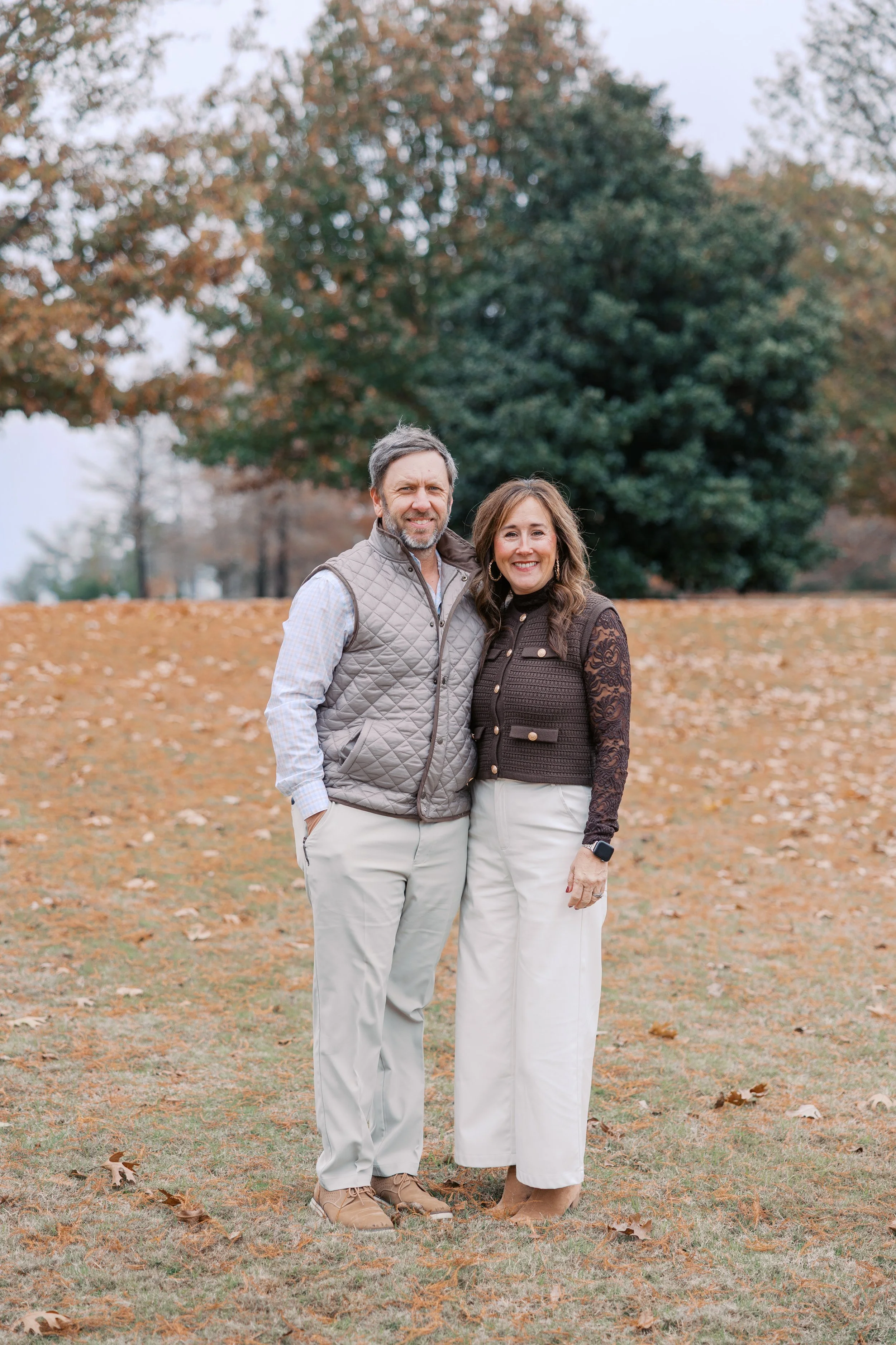 A middle-aged man and woman standing close together outdoors in fall, smiling at the camera, with trees and fallen leaves in the background.