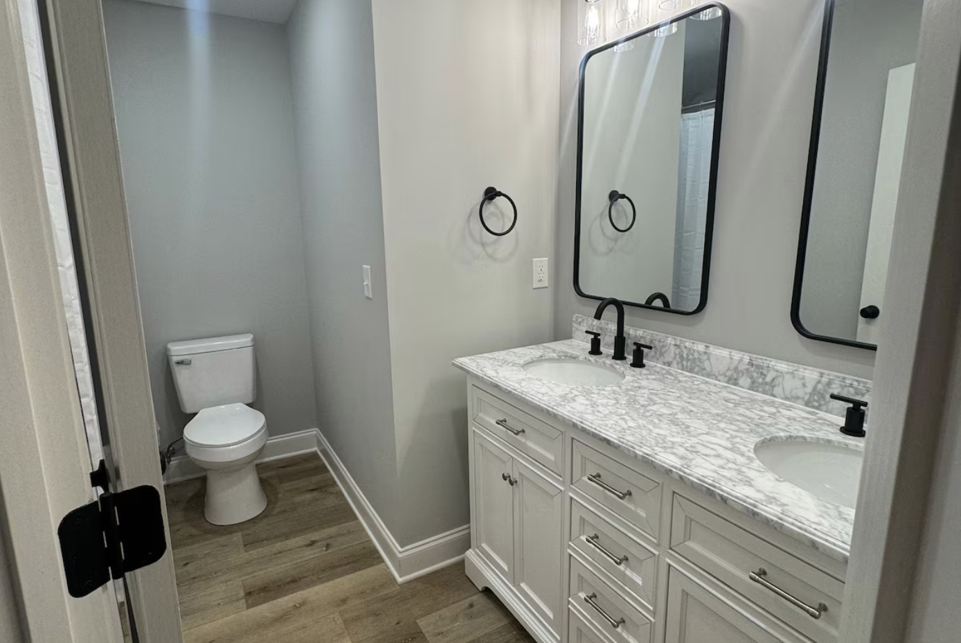 Bathroom with a double vanity sink with a marble countertop, two mirrors, and black fixtures; toilet in a separate small alcove; wood flooring; black towel rings; gray walls.