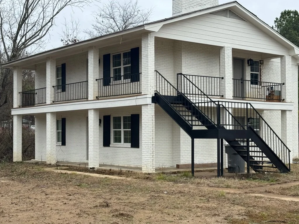A two-story white brick house with black shutters and black metal staircase leading to the second-floor porch, which has black railings and a porch swing. The yard is bare with sparse grass and dirt, and trees are visible in the background.