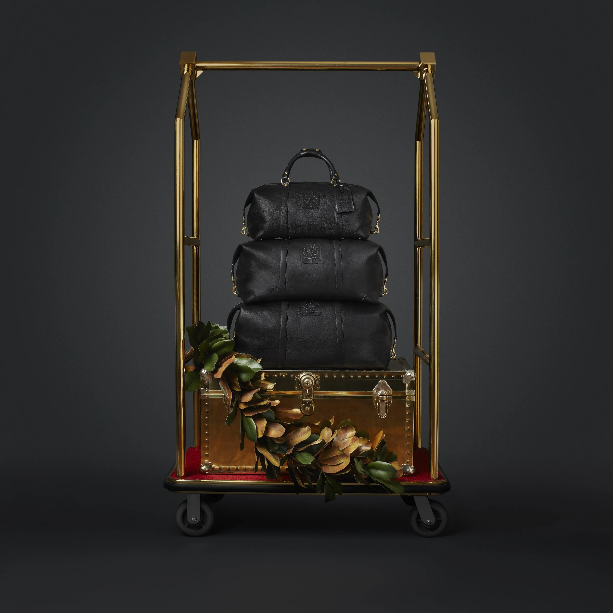 Ghurka leather Cavalier duffel bags stacked on brass luggage cart with magnolia leaves, luxury retail art direction by Alexander Mann