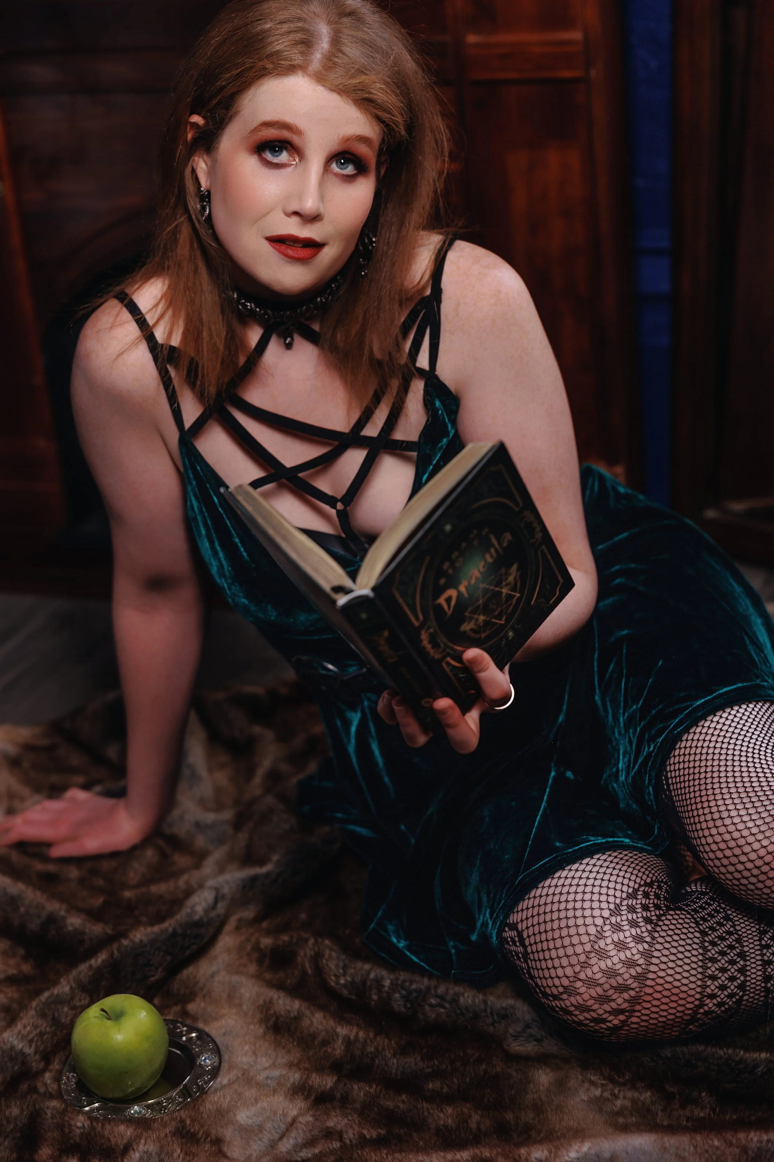 A tall redhead with blue eyes, freckles and red slipstick and eyeshadow looks into the camera while holding an open copy of Dracula by Bram Stoker. She's wearinga  green dress with strapy things over her cleavage and it's hot as fuck trust