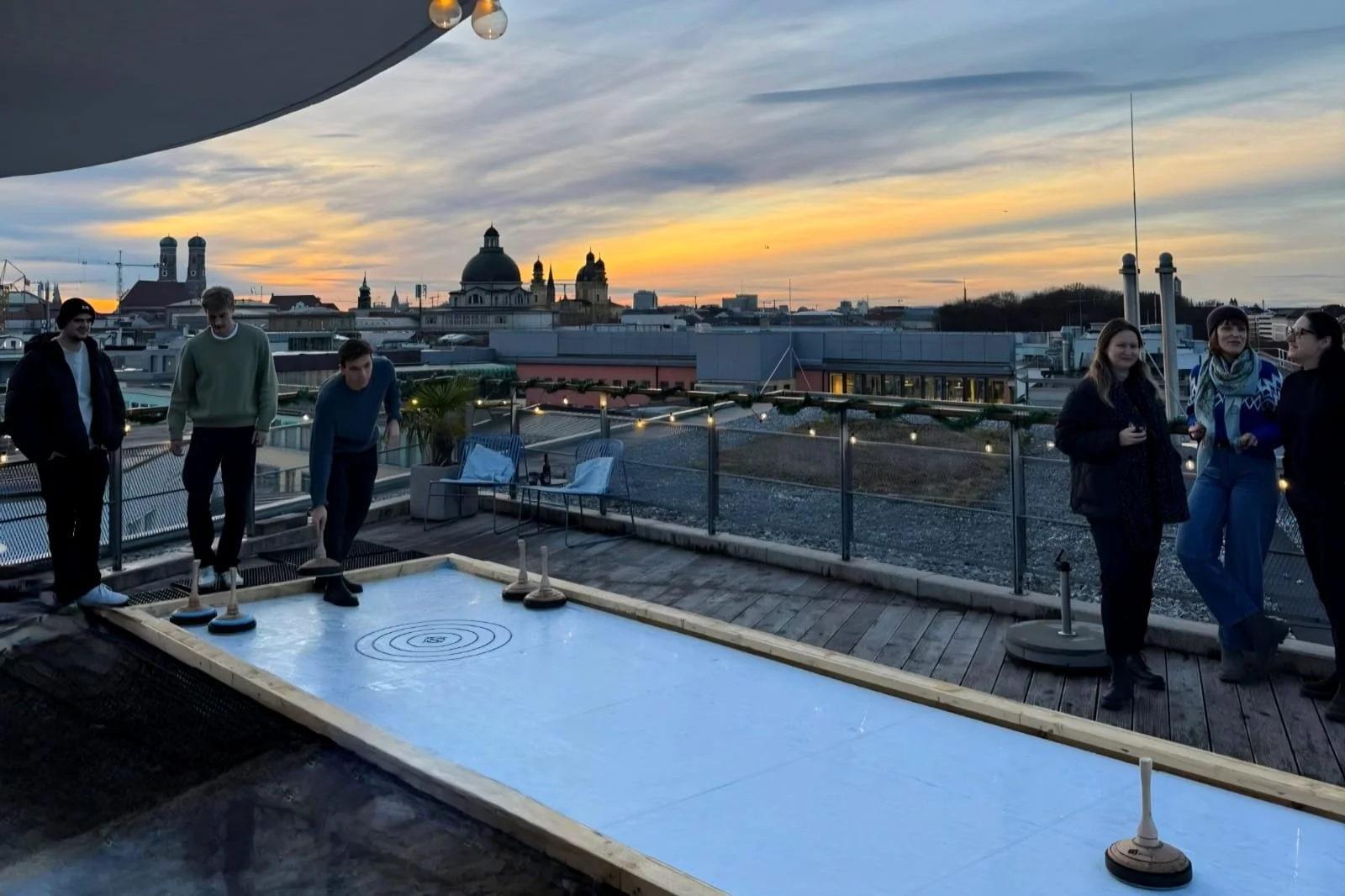 Eine Gruppe, die bei Sonnenuntergang auf einer Dachterrasse in München Eisstockschießen spielt.