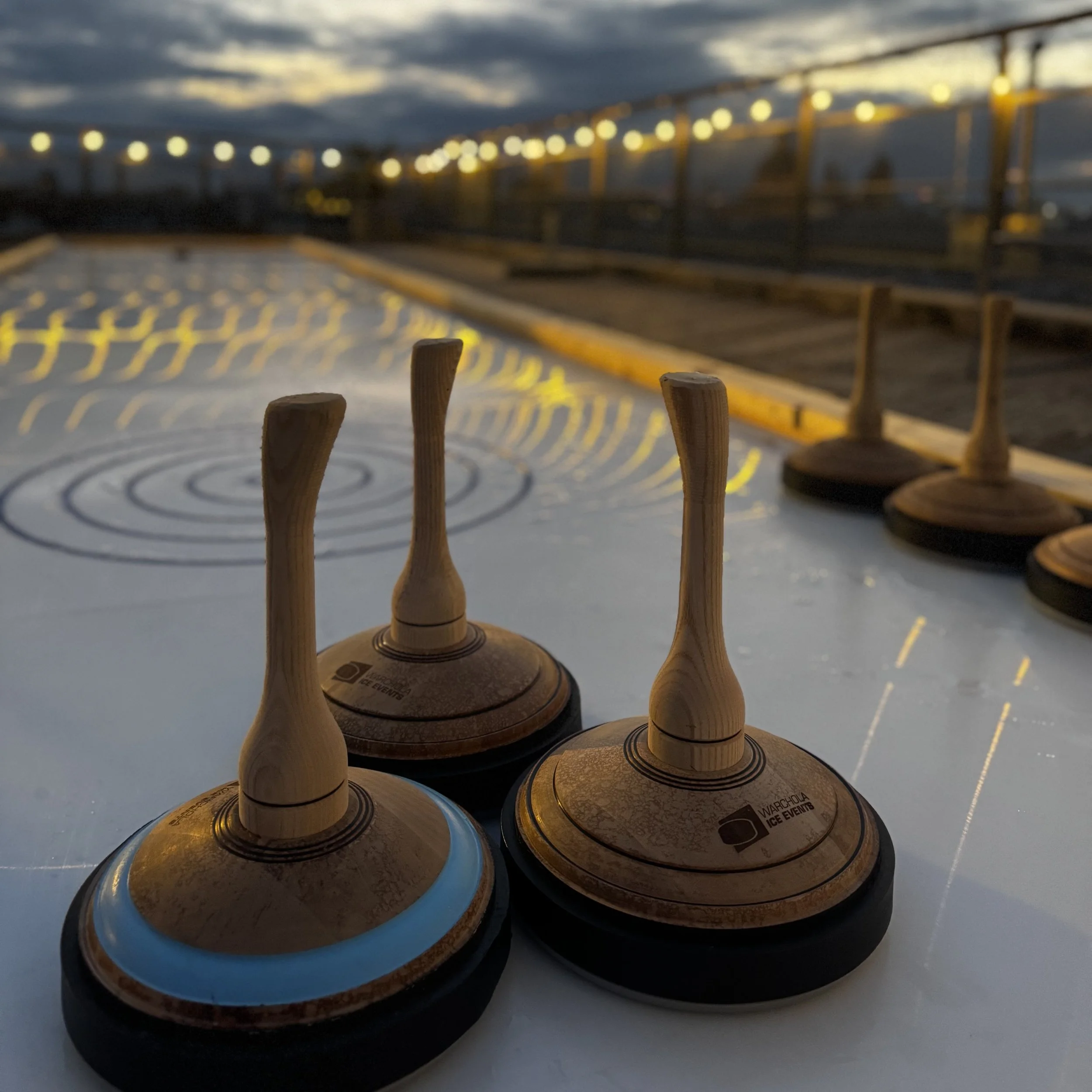 Ice hockey pucks and wooden curling stones on an outdoor ice rink at dusk with string lights in the background.