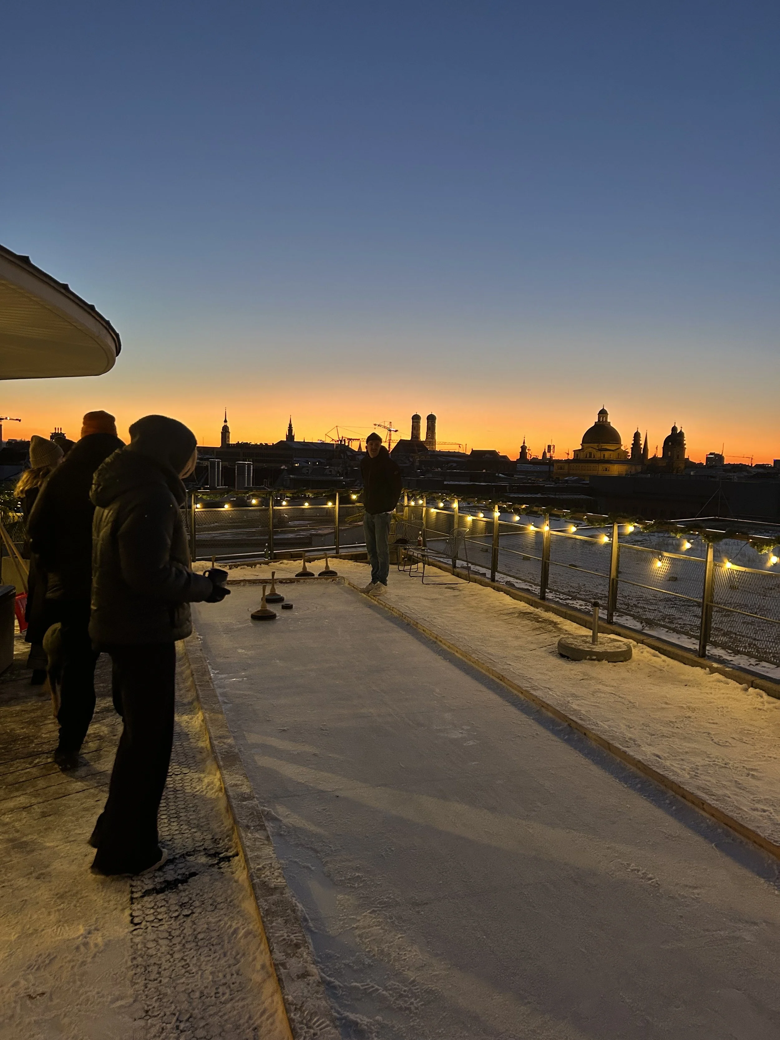 Eine Partie Eisstockschießen in München bei Schnee auf der Dachterrasse OutOfOffice Eisbach bei Sonnenuntergang.