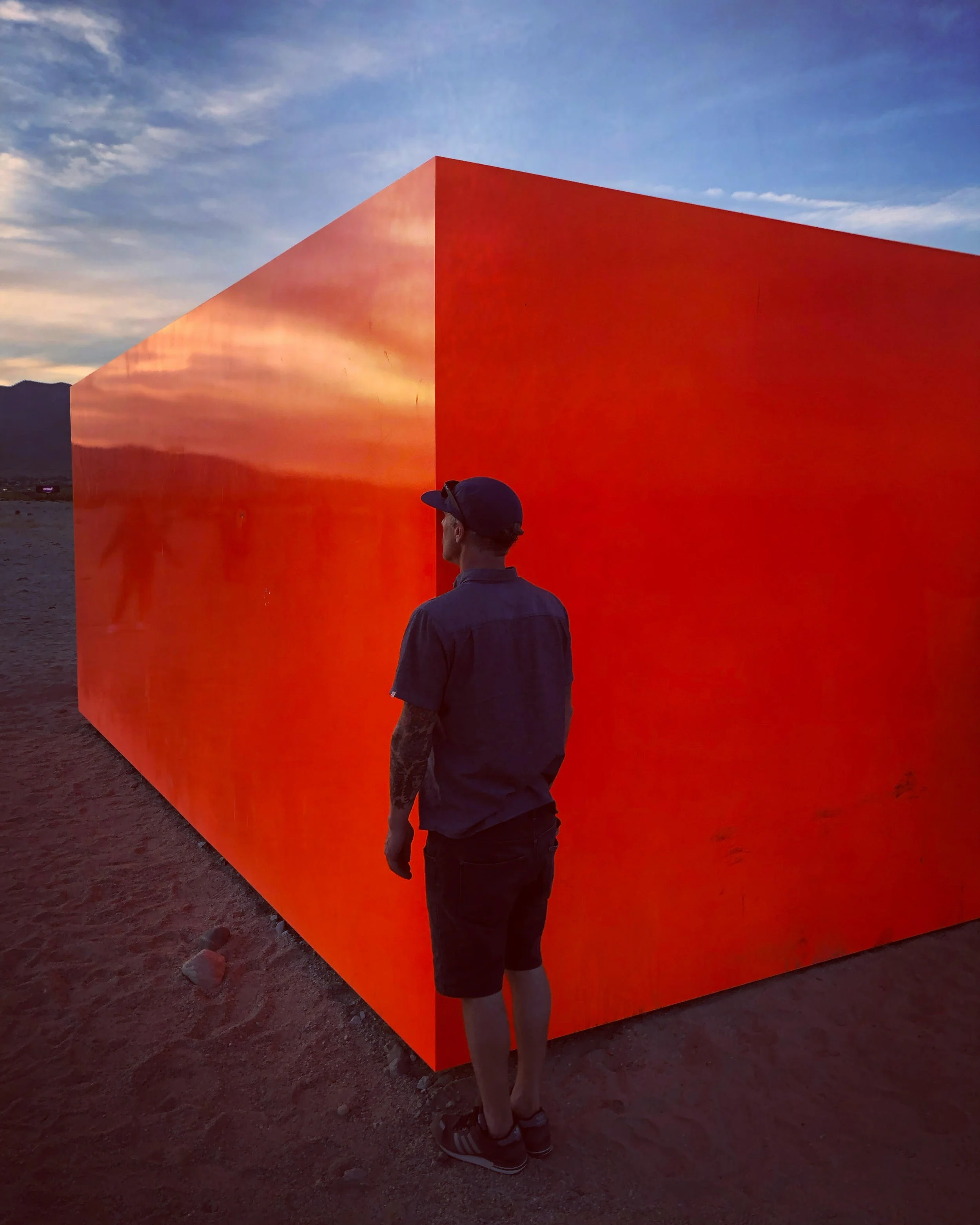 A man in a dark shirt, shorts, and a cap stands in front of a large, bright orange rectangular box outdoors at sunset with mountains and a partly cloudy sky in the background.