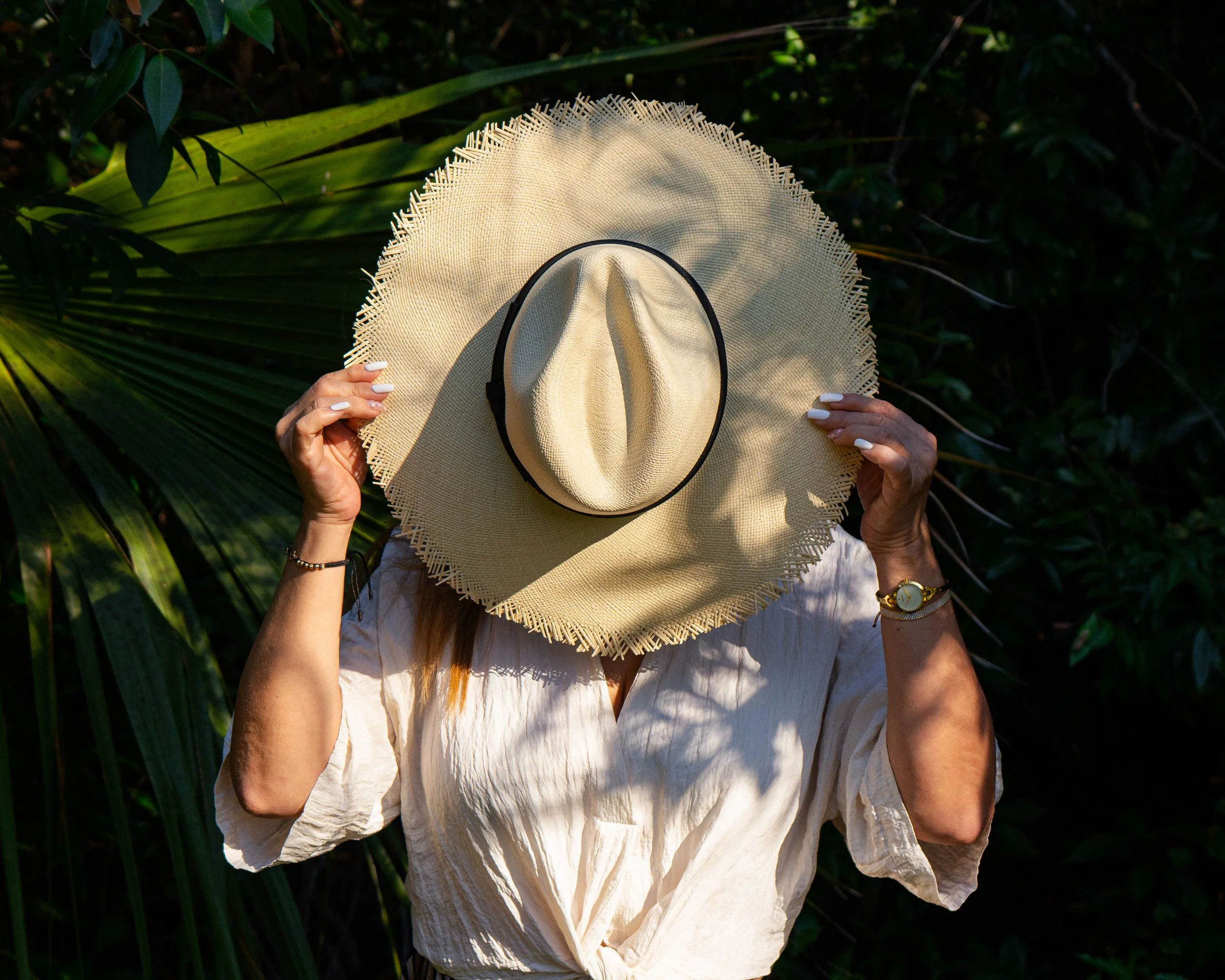 Person holding a large straw hat in front of their face, standing among green foliage.