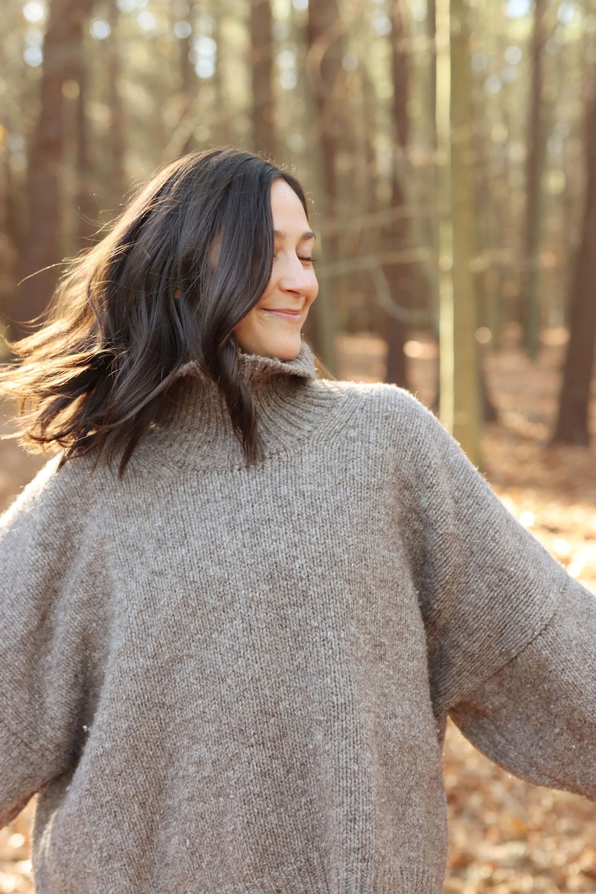 A woman with dark brown hair in loose waves, wearing a beige turtleneck sweater, standing in a forest with trees and sunlight filtering through.