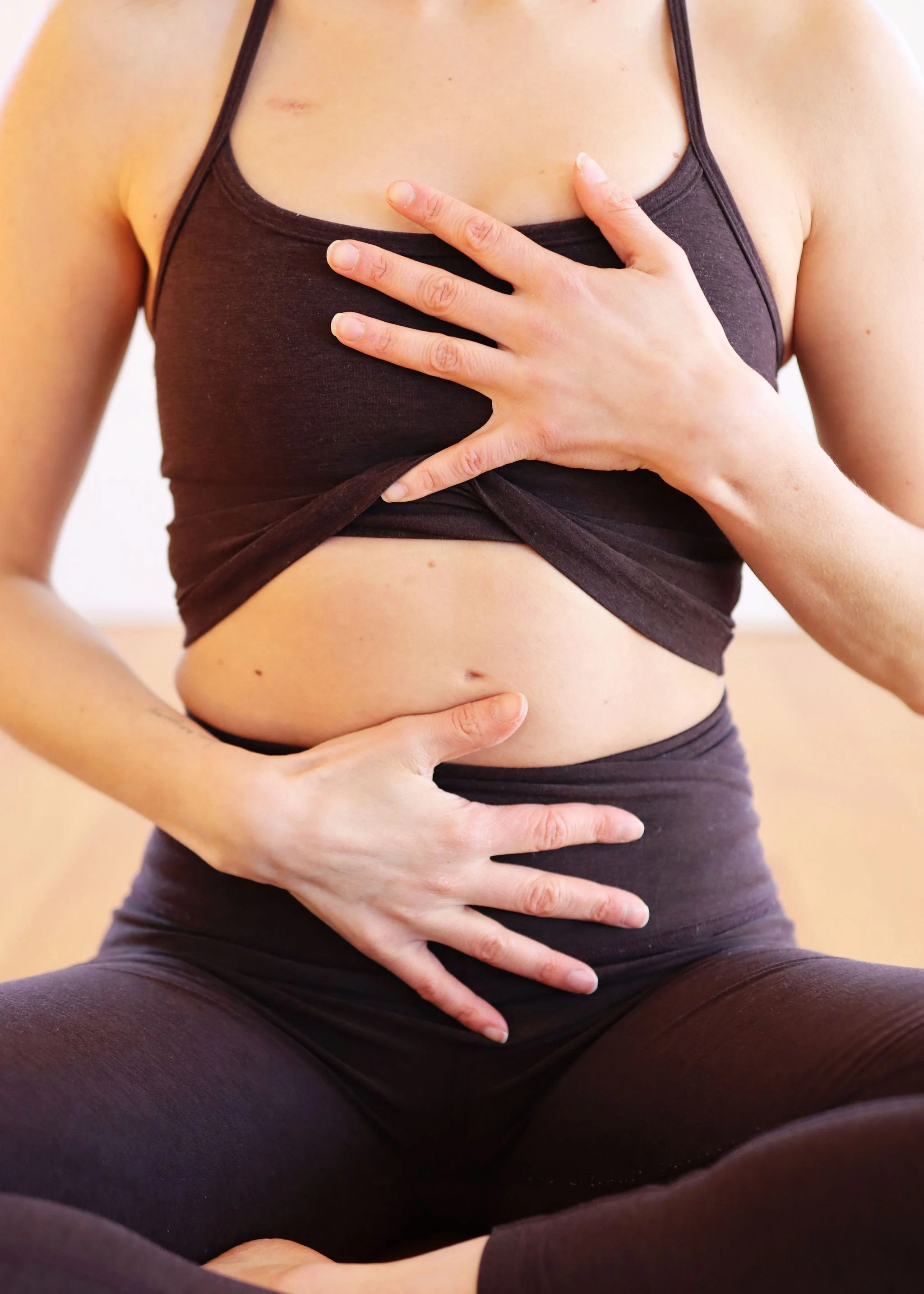 A person sitting cross-legged wearing a dark sports bra and leggings, with one hand on their chest and the other on their stomach, practicing yoga or meditation.