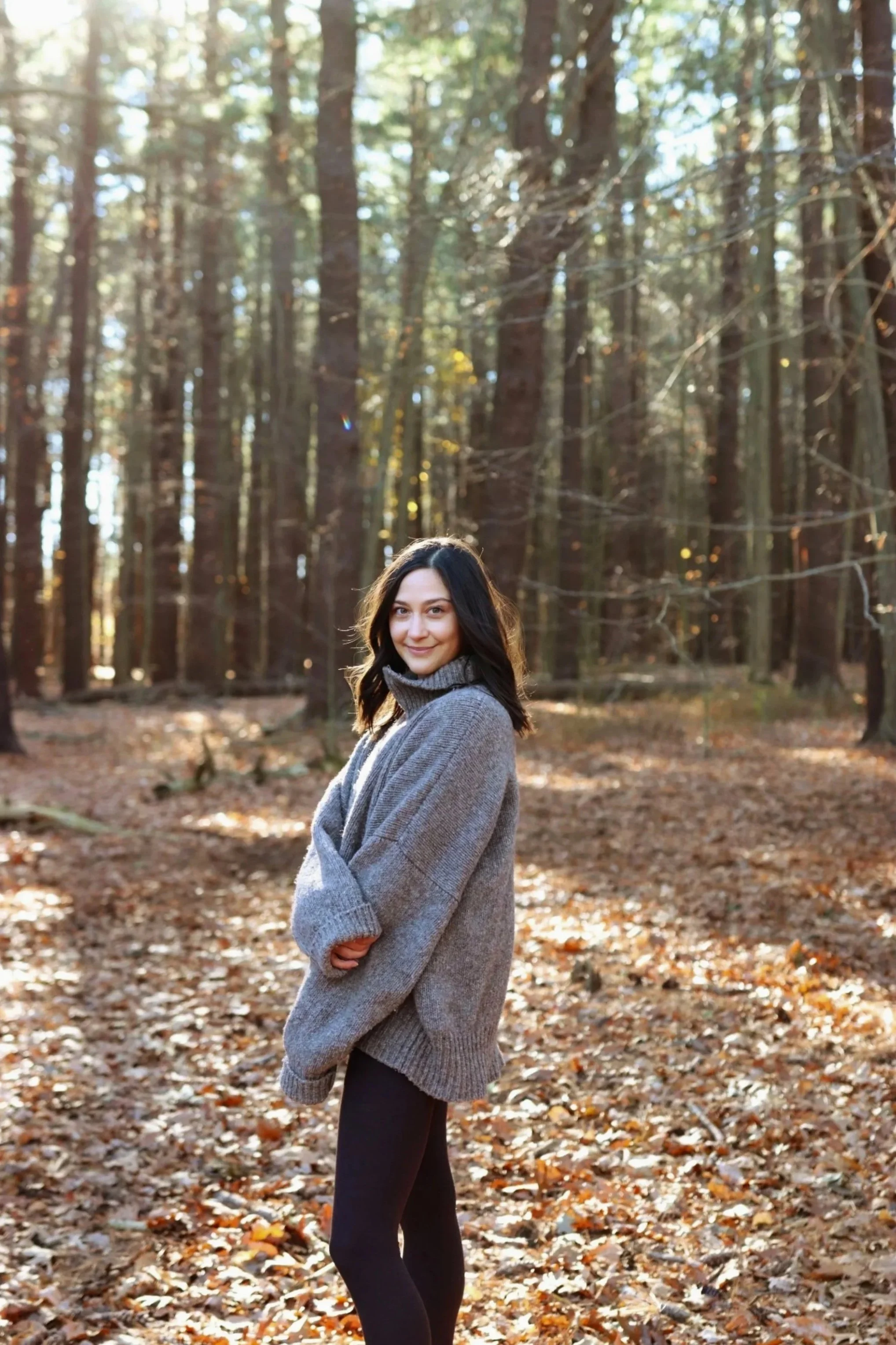 A woman in a gray sweater standing on a leaf-covered forest floor with tall trees in the background during daylight.