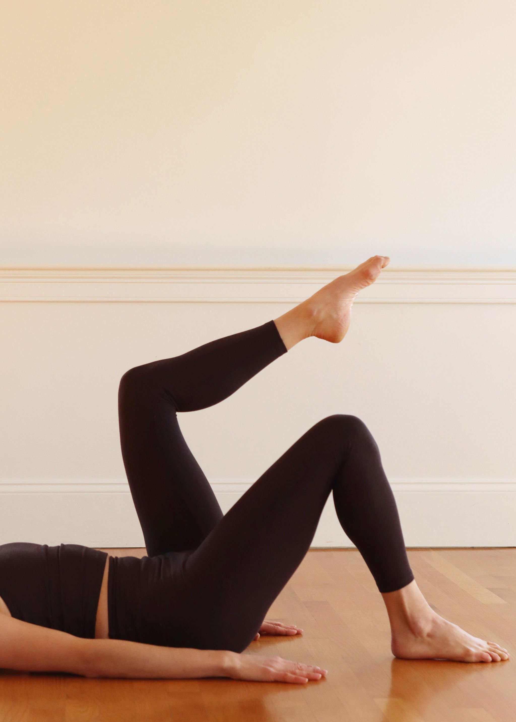 A person lying on the floor in a yoga pose, with one leg extended upward and the other bent, wearing black leggings and a black top, on a wooden floor with a plain light-colored wall in the background.