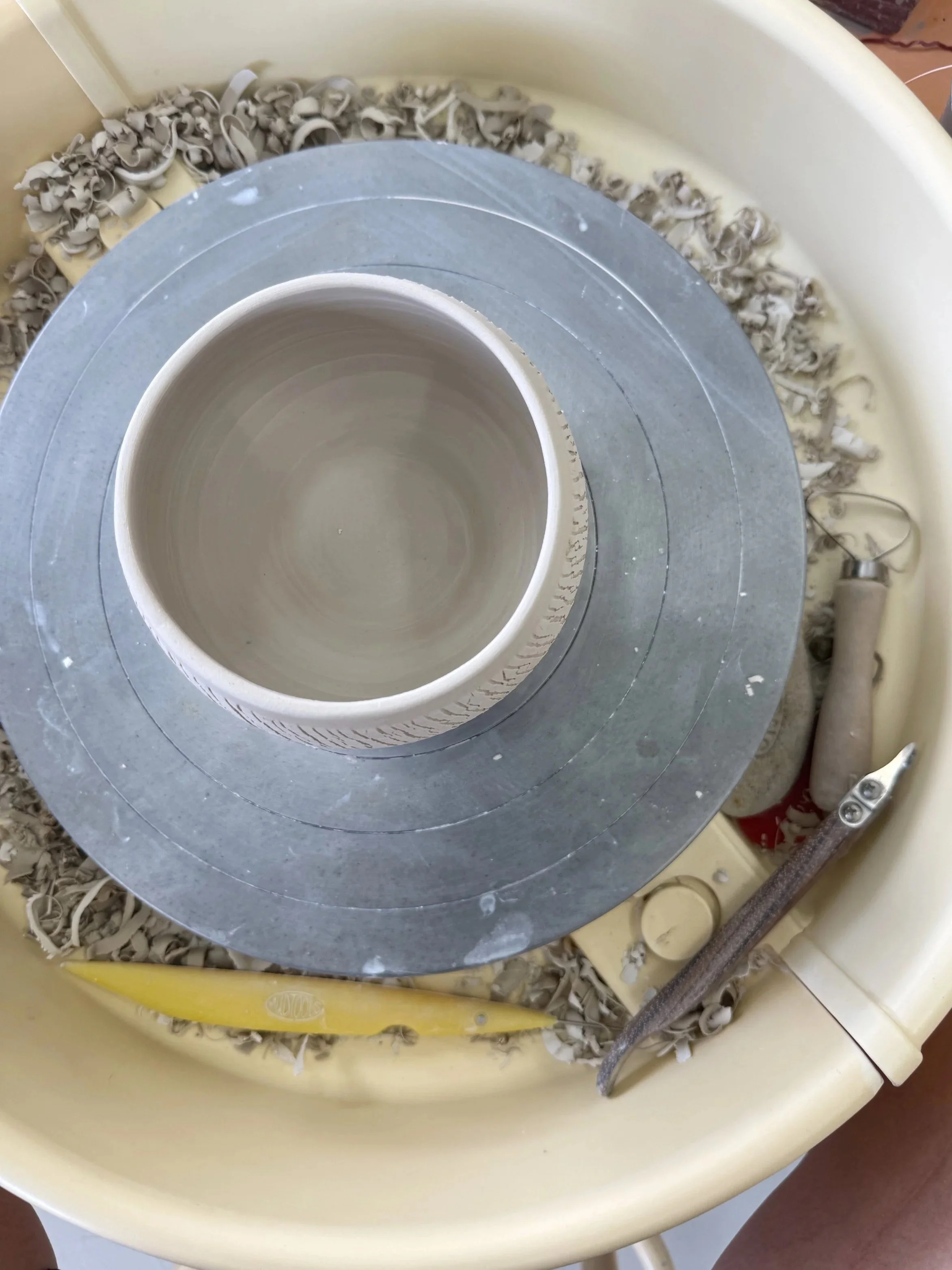 A pottery wheel with an unshaped clay vessel in the center, surrounded by clay shavings, with tools such as a metal rib and a small sponge nearby in a pottery studio.