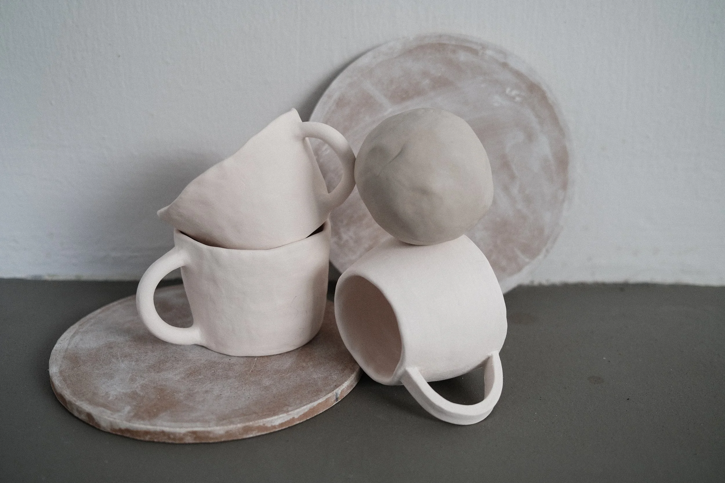 Stacked white ceramic cups, one with a ball of clay on top, set on a rustic round clay plate with a smaller plate in the background.