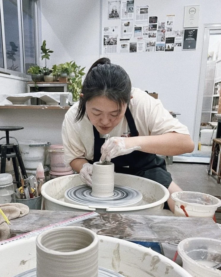A person working on a pottery wheel shaping a ceramic piece in a pottery studio. There are various supplies and a wall with pictures and articles behind them.
