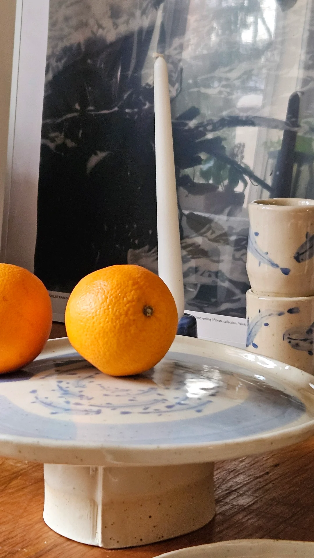 Kaksi appelsiinia ja kaksi keramiikkamukia pöydällä, taustalla valkoinen kynttilä ja taulu tai kuva.