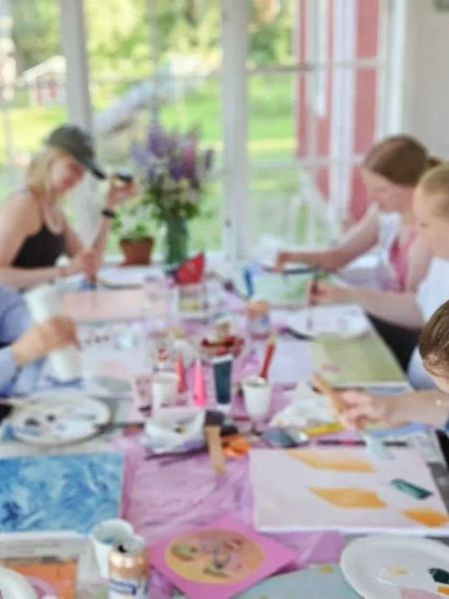 Sisätiloissa kuva, jossa useita ihmisiä istuu pöydän ympärillä. Pöydällä on värejä, paperia ja maaleja, ja ihmiset näyttävät harjoittelevan taidetta tai askartelua. Taustalla on suuret ikkunat, joiden kautta näkyy vihreää nurmikkoa ja puita.