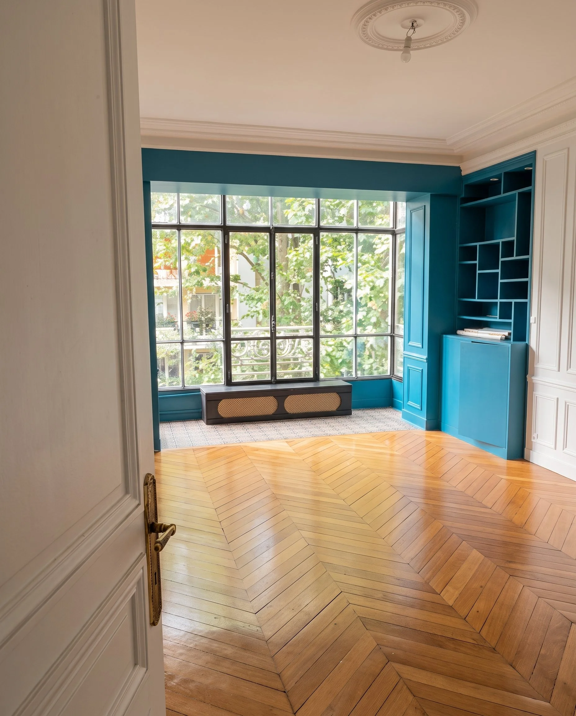 Pièce lumineuse d'un appartement parisien avec un bow-window donnant sur des arbres, parquet point de hongrie, bibliothèque intégrée bleue, et un plafond avec moulures