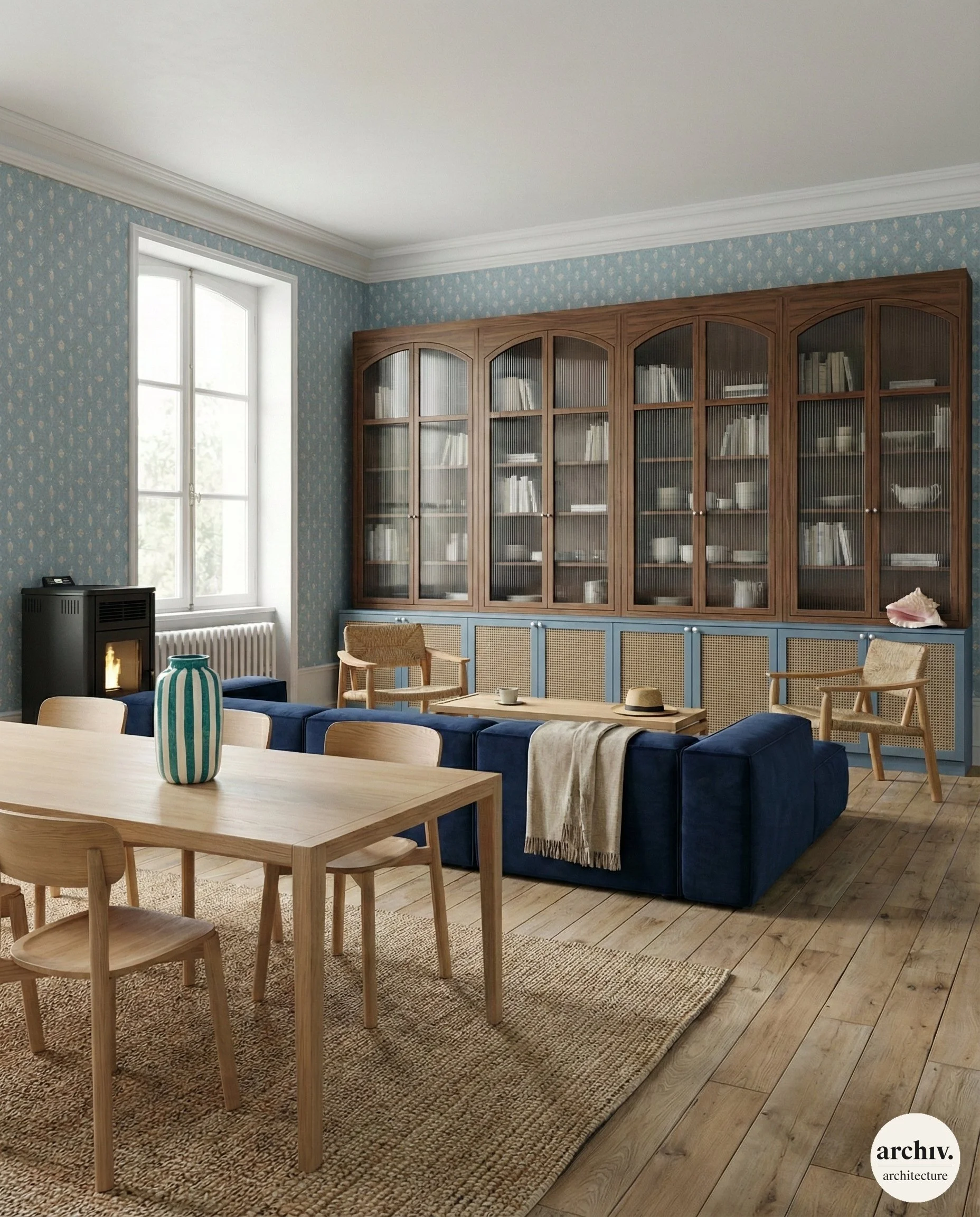 Living room with a large blue sectional sofa, wooden coffee table, and beige chairs against a wall with a wooden bookshelf. There is a window with natural light, and a fireplace. The room has wooden flooring and patterned wallpaper.  Bretagne.