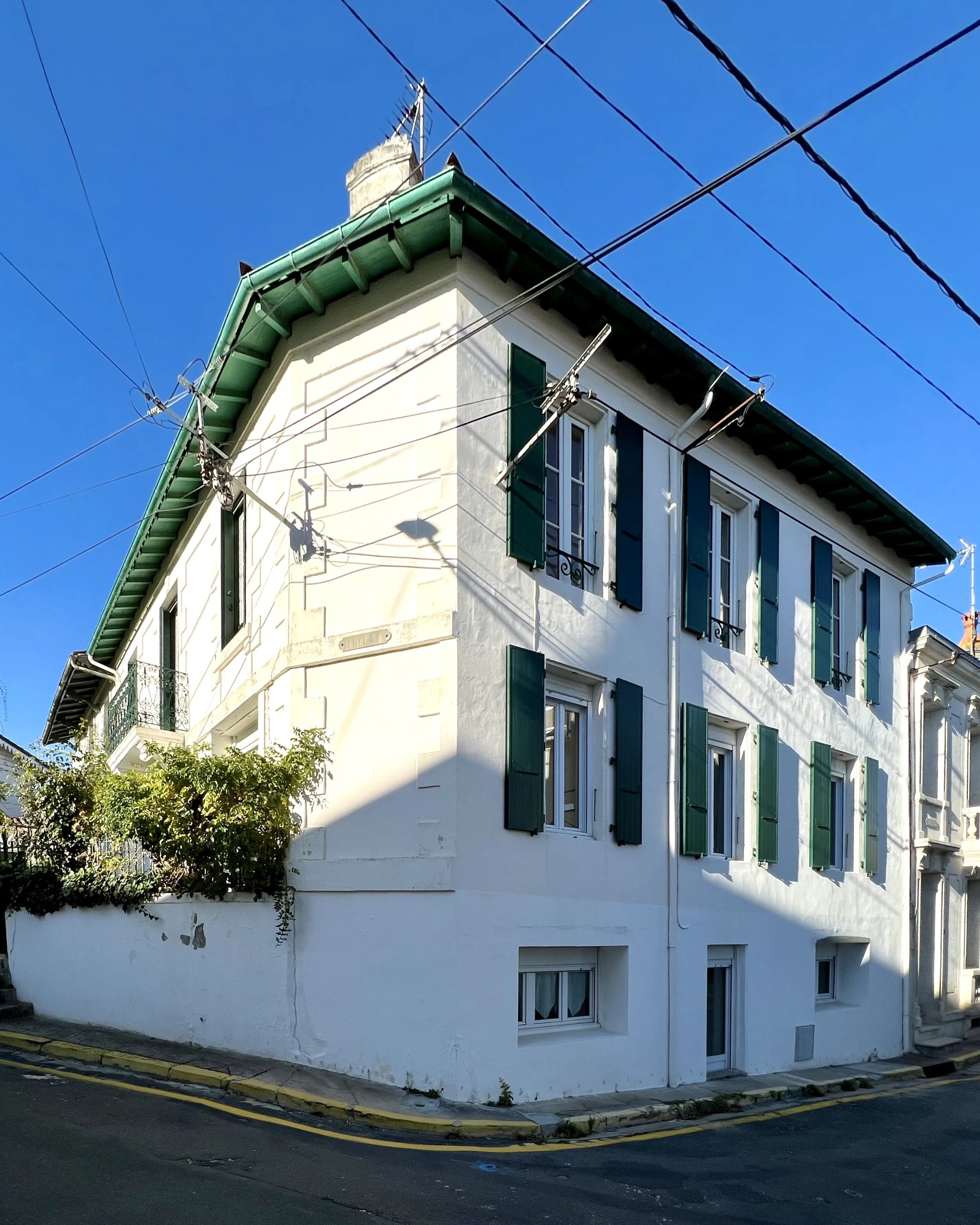 Maison charentaise blanche avec fenêtres à volets verts, située au coin de la rue.