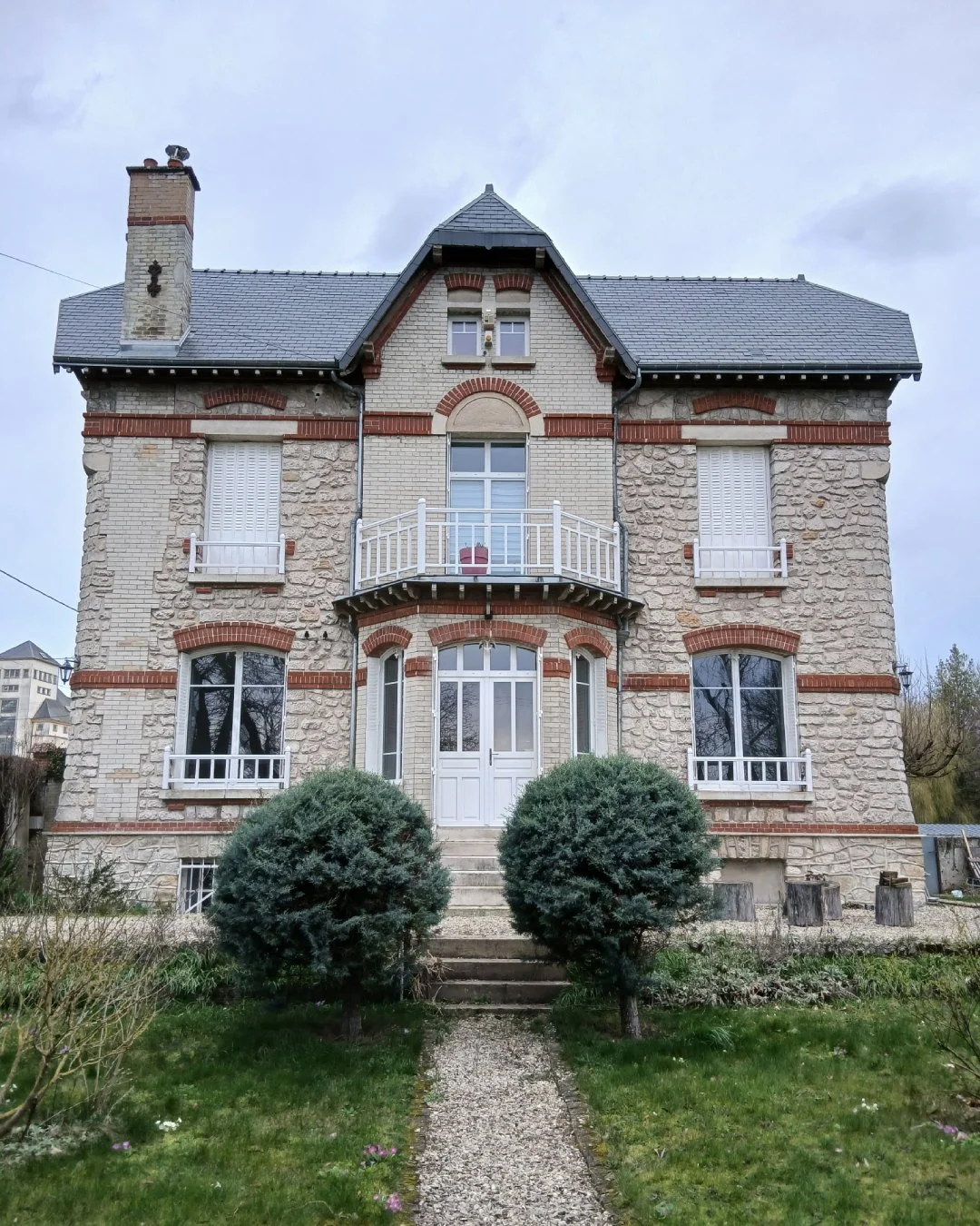 Maison de maître en pierre avec toit en ardoise, fenêtres avec persiennes, balcon.