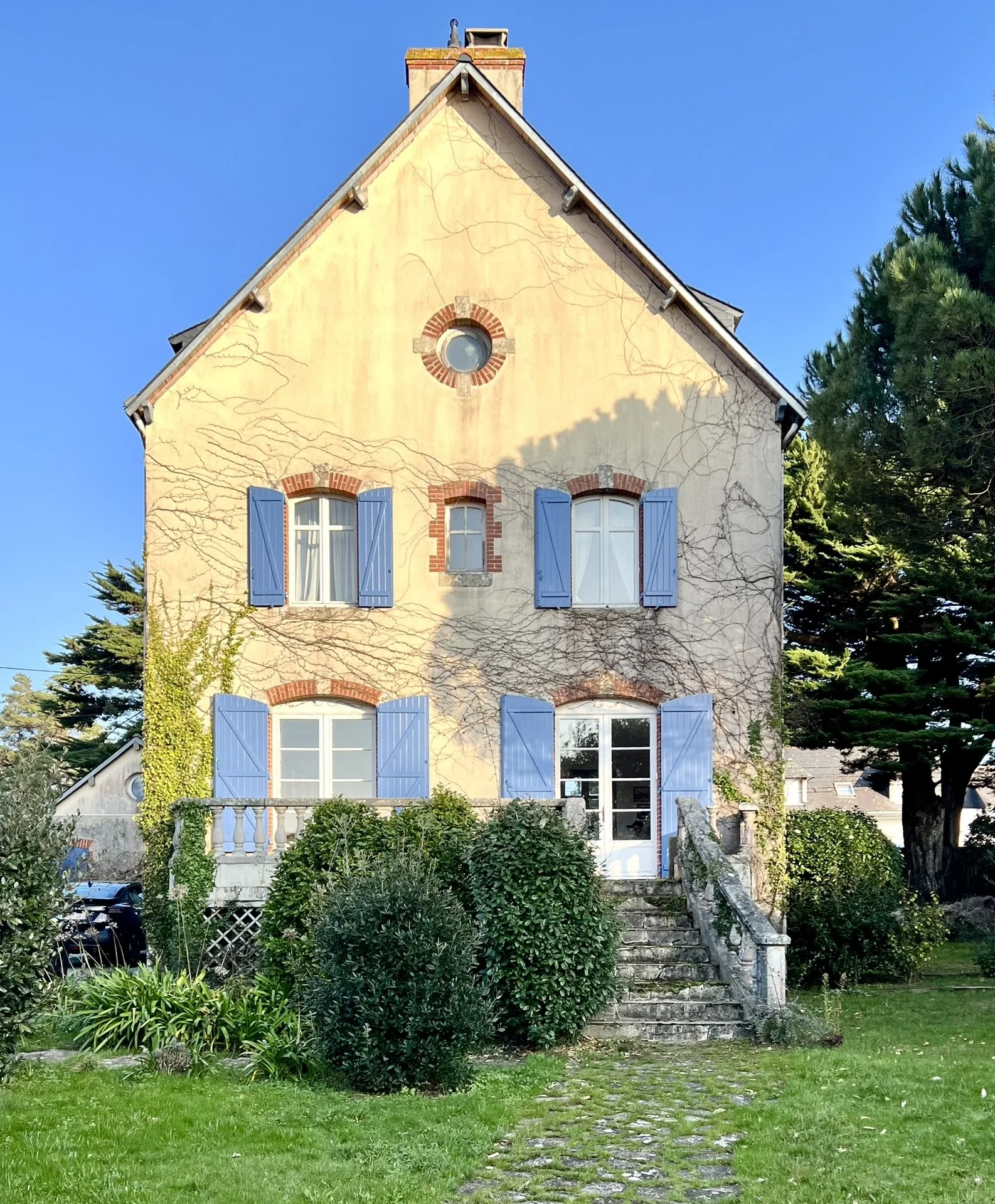 Vue d'une maison breton avec enduit décrépi et volets bleus, entourée d'un jardin verdoyant, escalier menant à la porte d'entrée.