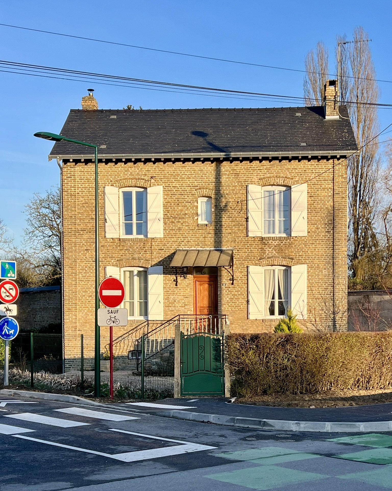 Maison en briques ocres avec fenêtres à volets battants blancs, entourée d'un jardin clôturé, à l'angle de rues.
