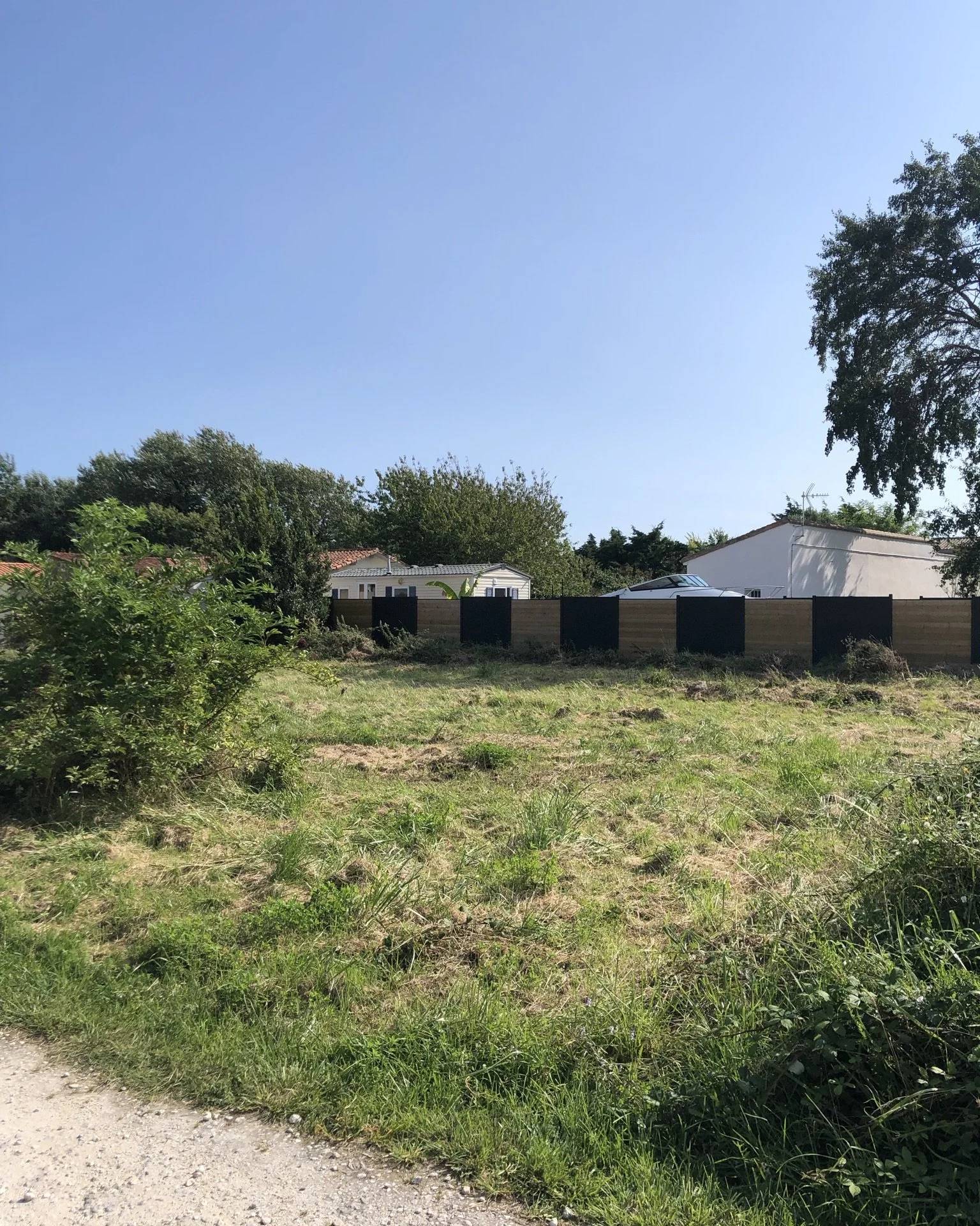 Jardin en Charente maritime avec herbe, buissons et arbres, clôture en bois, maisons avec toits en tuiles.