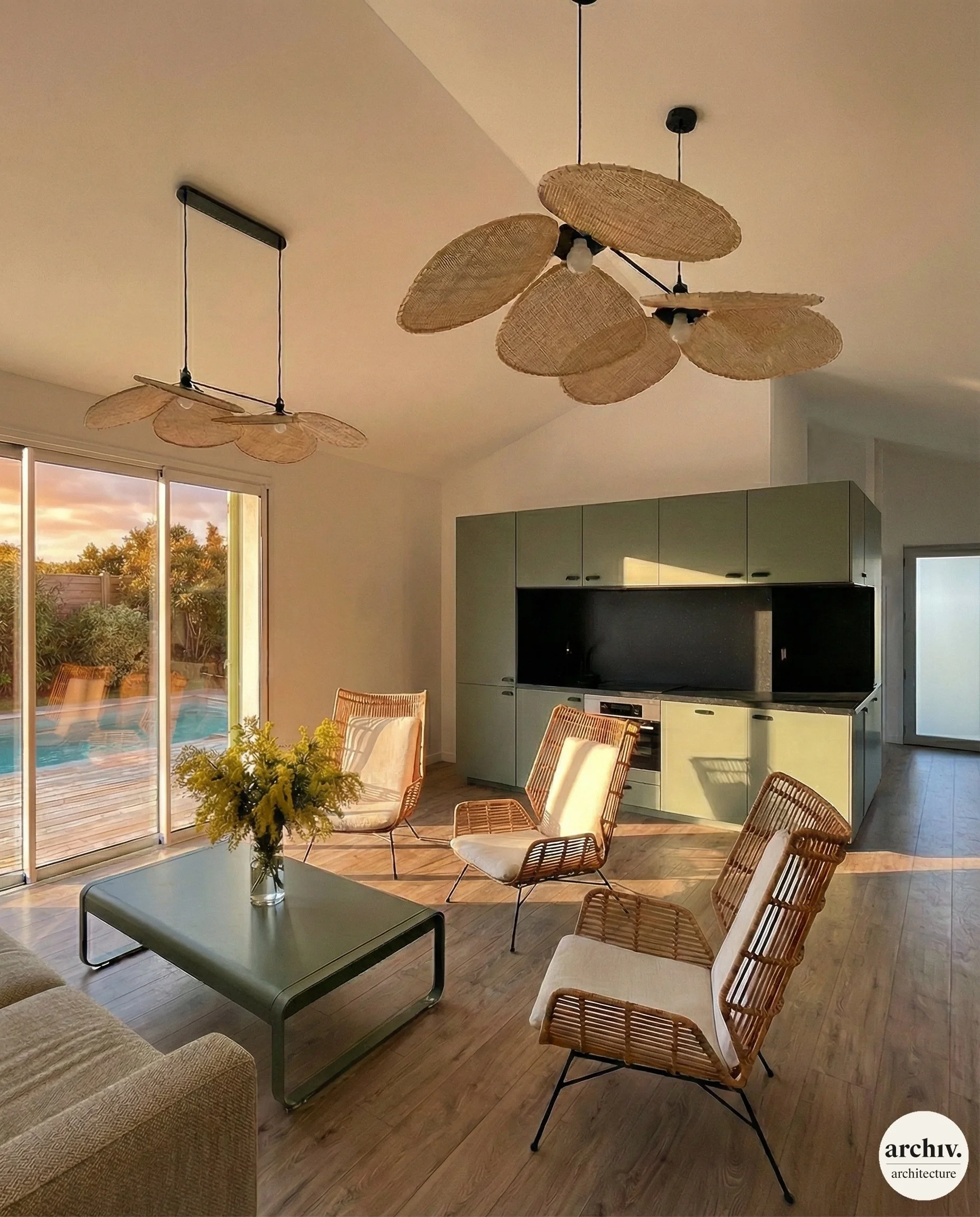 Interior of a contemprorary living room with a green kitchen, rattan chairs with white cushions, a coffee table, and two ceiling lamps with woven shades. Large sliding glass doors show an outdoor pool and garden.  Royan.