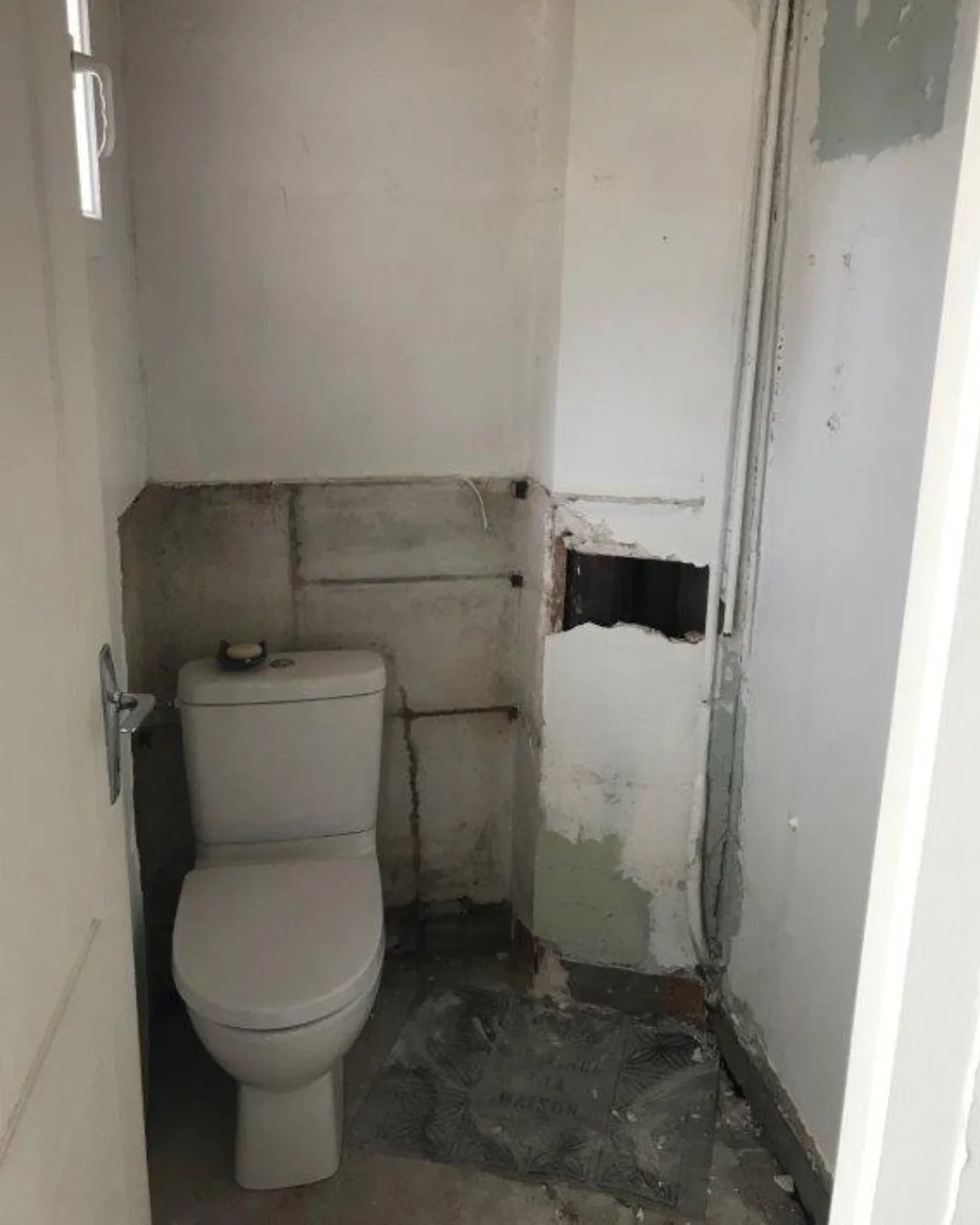 Salle de bain en rénovation avec wc blanc, murs en cours de travaux, tuyaux et câbles apparents, sol en carrelage abîmé.