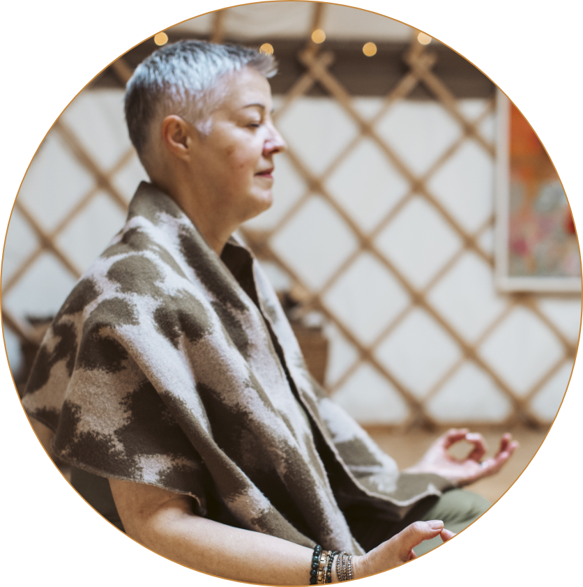 A person practicing meditation indoors, sitting cross-legged with eyes closed, wearing a patterned shawl, and surrounded by warm lighting.
