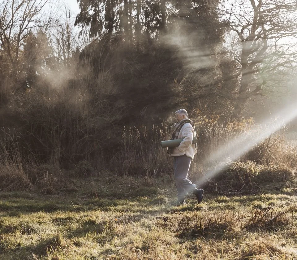 A man hiking outdoors in a grassy area with trees, carrying a rolled-up mat, with sunlight filtering through the trees.