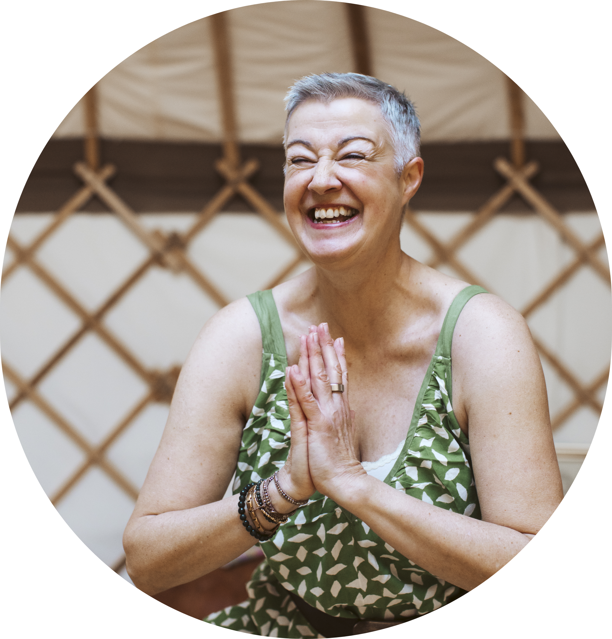 A joyful woman with short gray hair, smiling with her eyes closed, wearing a green patterned sleeveless top and multiple bracelets on her left wrist, standing inside a yurt or similar structure with a lattice pattern in the background.