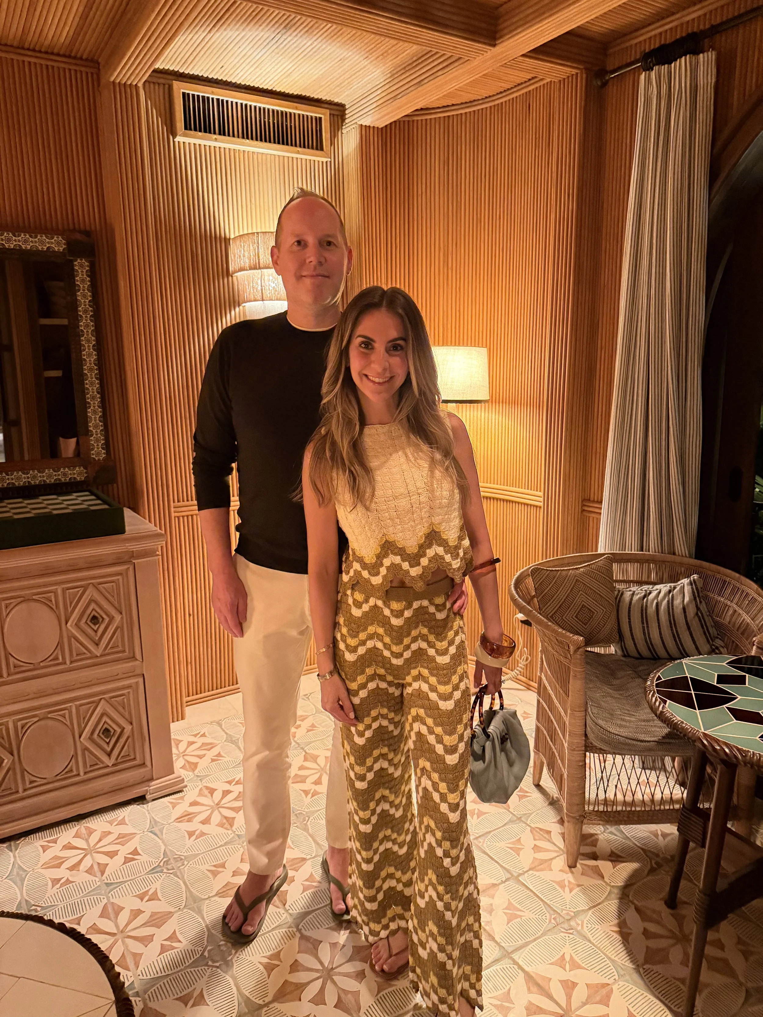 A man and woman standing inside a warmly lit room with wooden paneled walls, patterned floor tiles, and wicker furniture. The woman is wearing a patterned jumpsuit and holding a bag, while the man is dressed in a black top and white pants.
