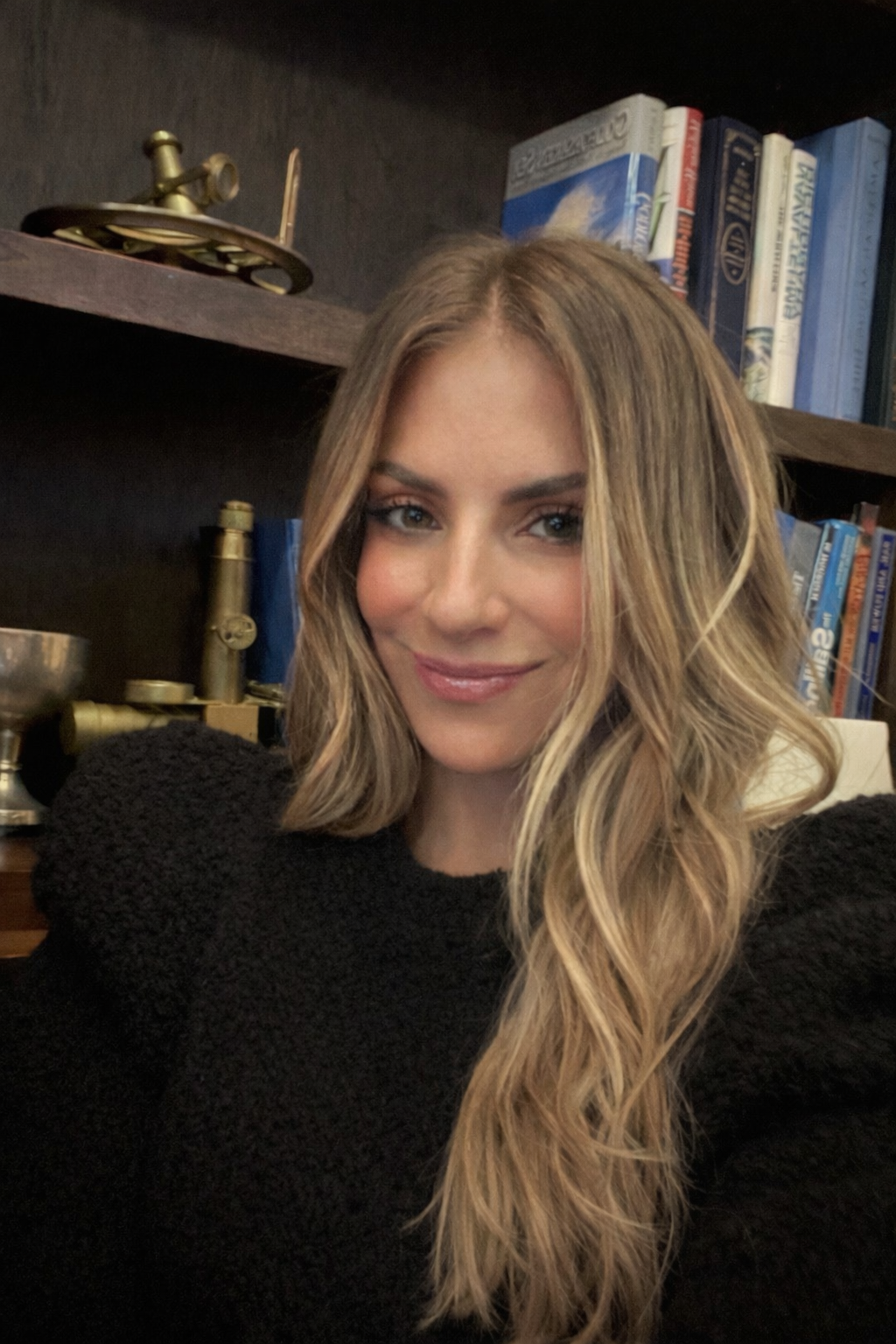 A woman with long wavy blonde hair and light makeup smiling at the camera, wearing a black sweater, in front of a bookshelf with books and decorative items.