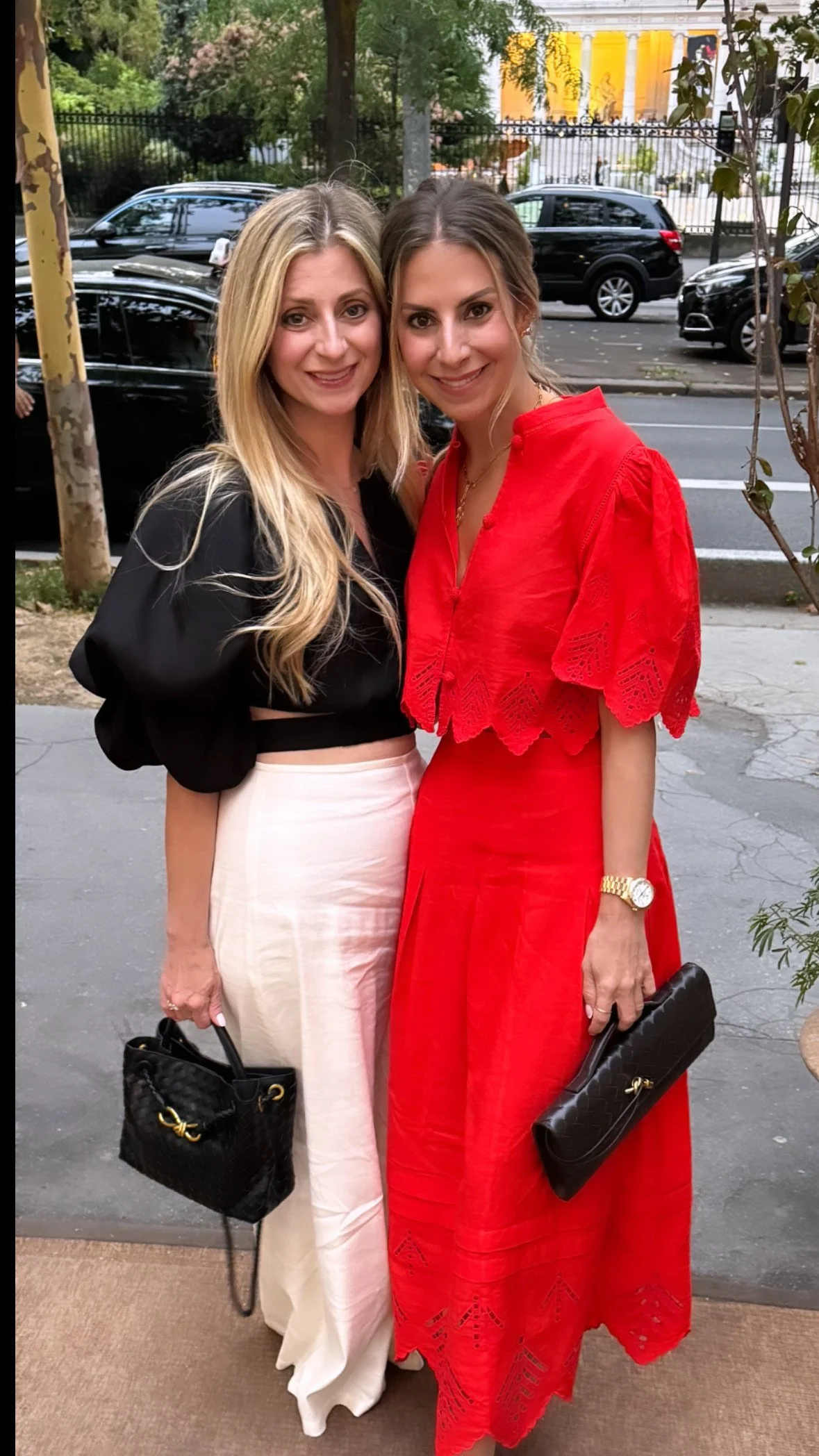 Two women standing outside, smiling, holding black handbags, with a street and parked cars in the background.