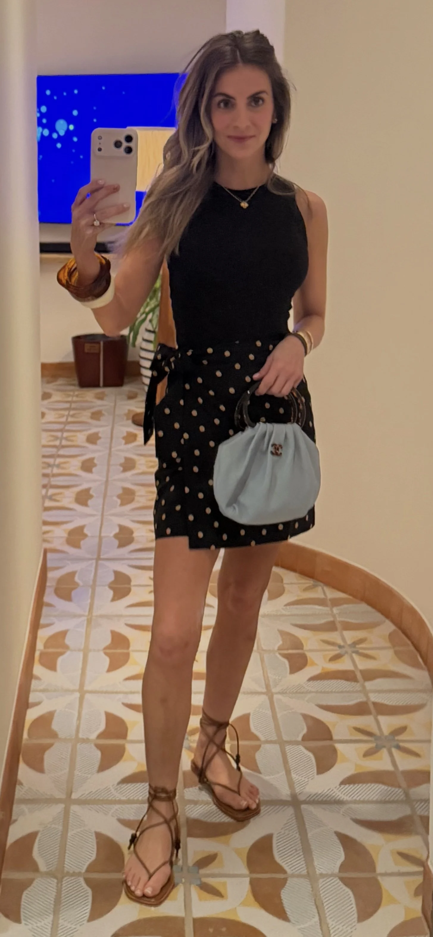 A young woman taking a mirror selfie in a hallway. She is wearing a black sleeveless top, a black skirt with white polka dots, and brown lace-up sandals. She holds a small light-colored handbag and has accessories including bracelets and a necklace. Her hair is styled in loose waves.