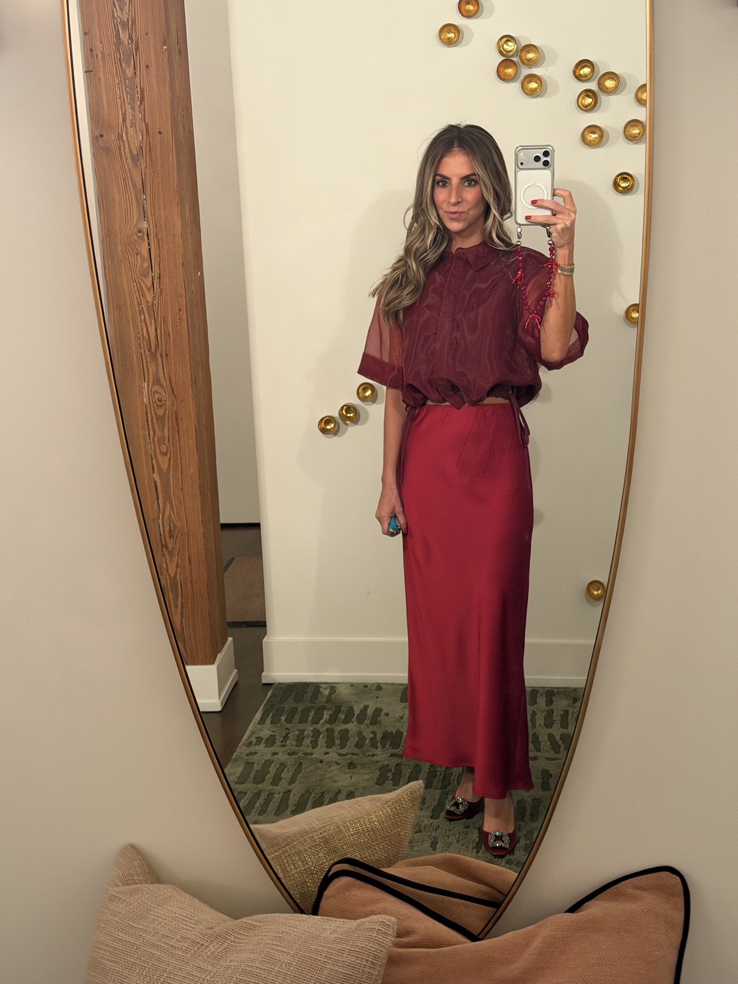 A woman taking a mirror selfie in a room with a beige wall decorated with small gold balls. She is wearing a burgundy blouse and a matching burgundy skirt, with black shoes adorned with embellishments.