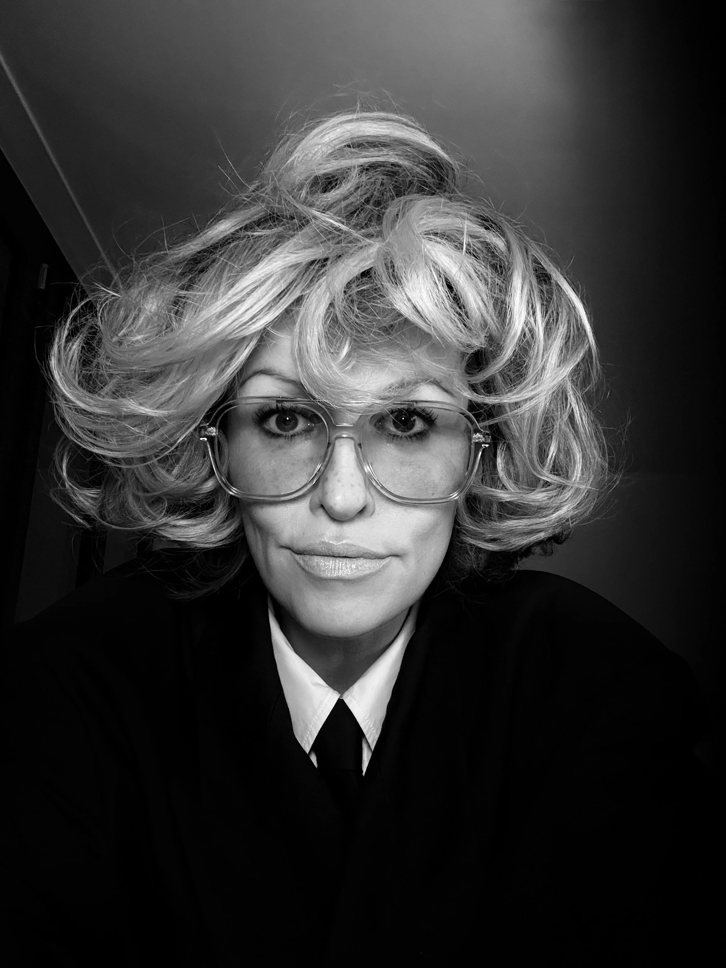 A woman with voluminous, curly blonde hair and glasses, wearing a black blazer and white shirt, looking directly at the camera in black and white.