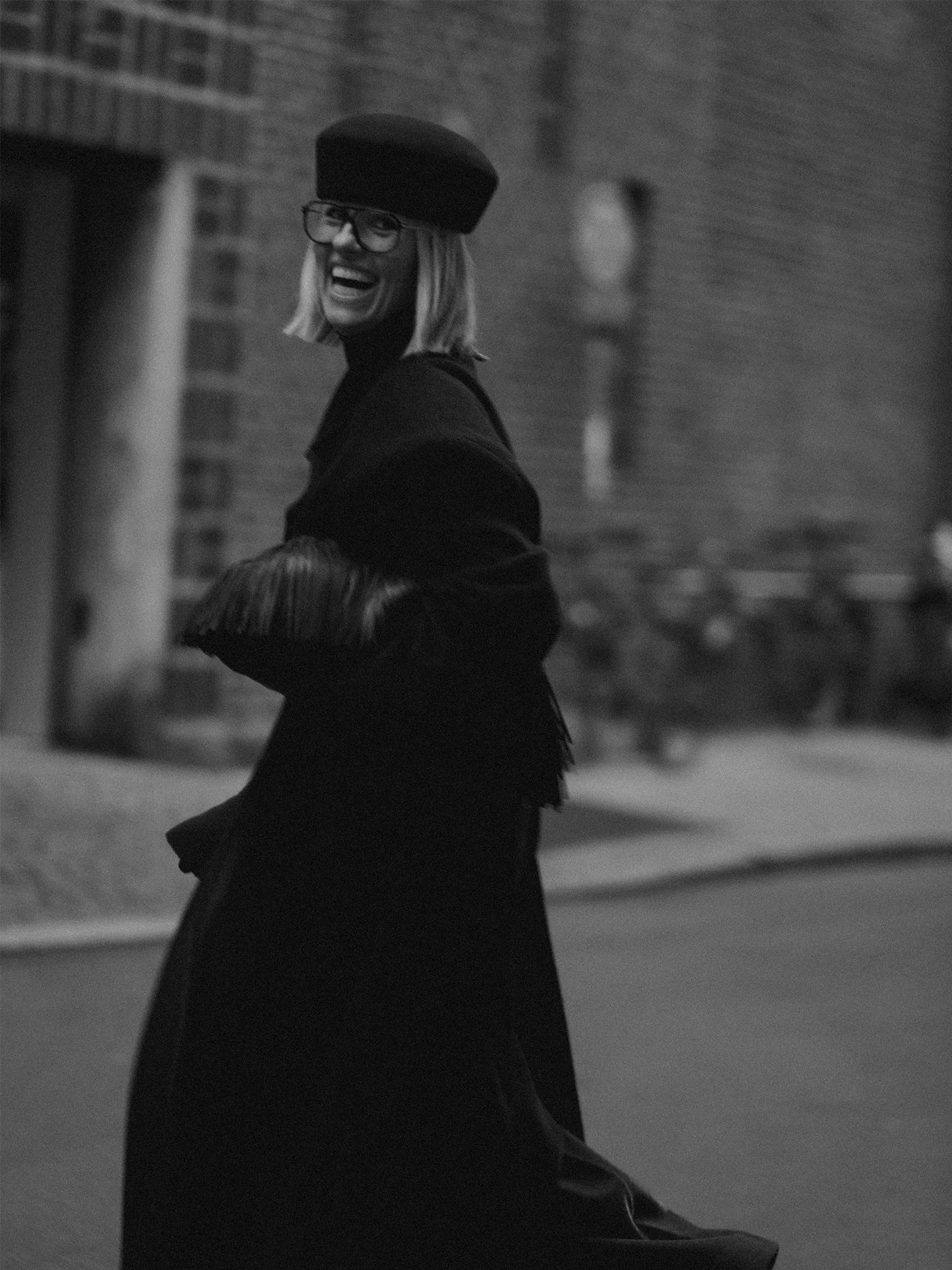 A woman with glasses and a hat, smiling and laughing outdoors at night, with a brick building in the background.