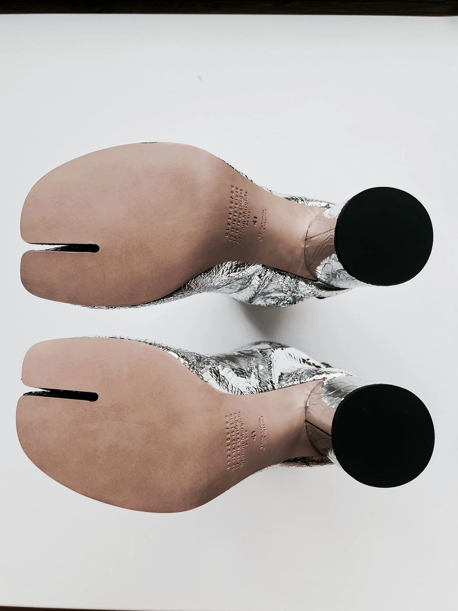 Two high heeled Maison Margiela Tabi Boots in broken mirror leather, viewed from the bottom on a white surface.