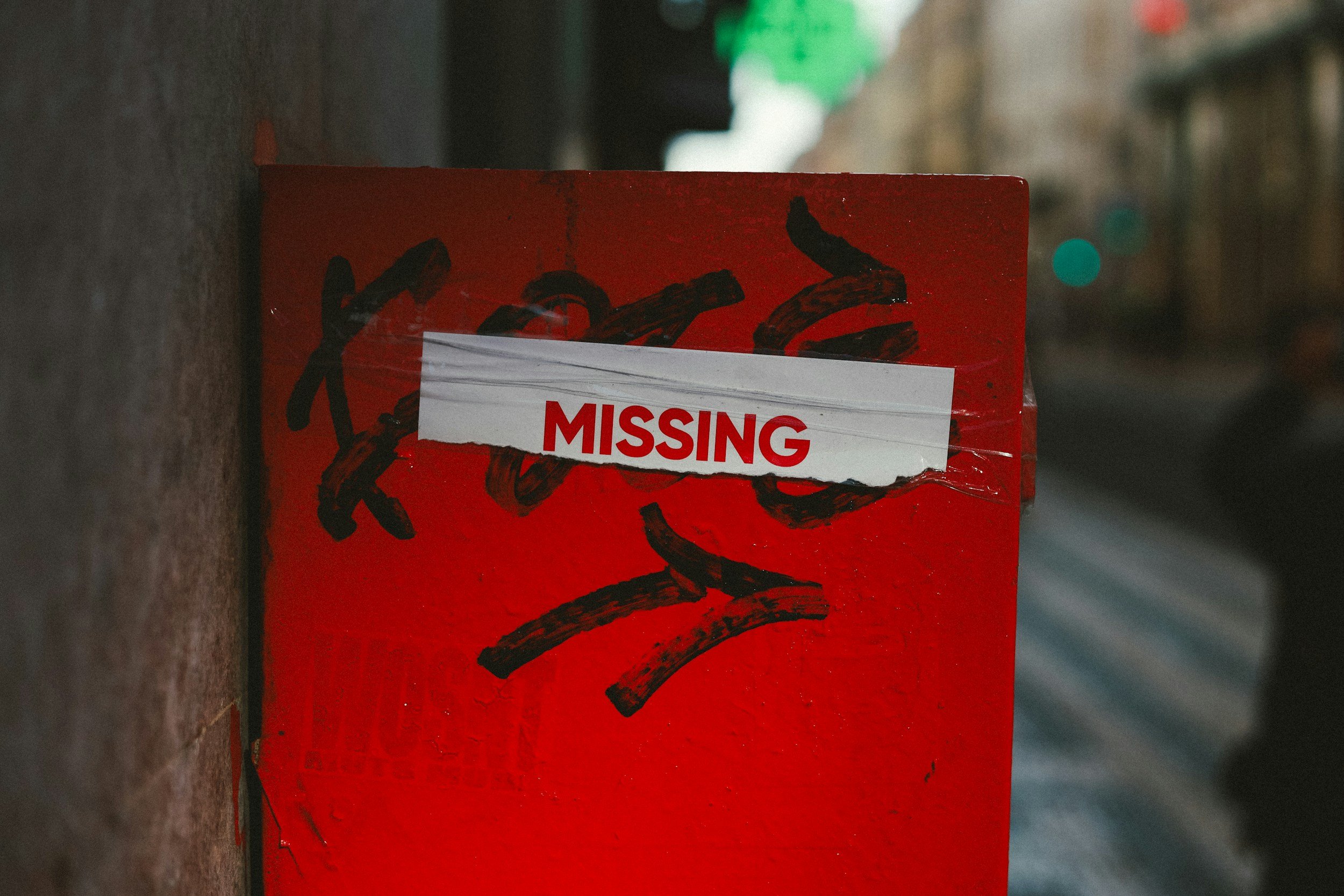 Red box on side of street with a white paper label that says 'MISSING' in red text, partially obscured by clear tape, and black graffiti on the box.