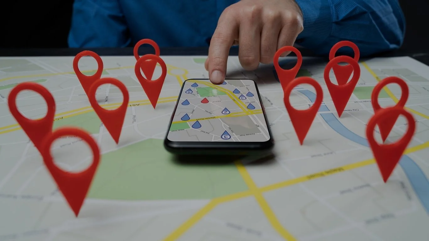 Person pointing to a smartphone displaying a map on a table with a printed map underneath, surrounded by several red map markers.