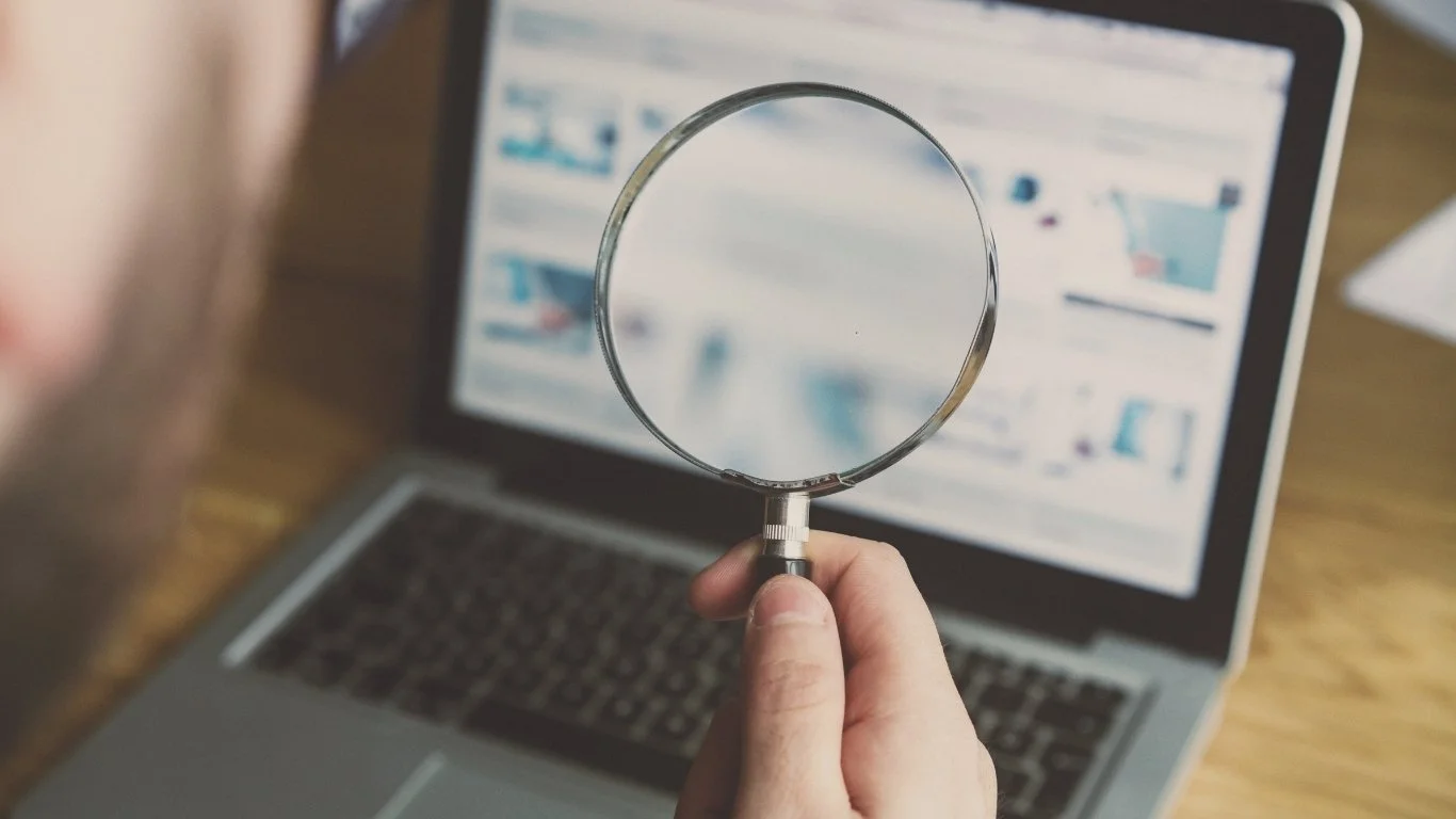 A hand holding a magnifying glass in front of a laptop displaying blurred search results or images.