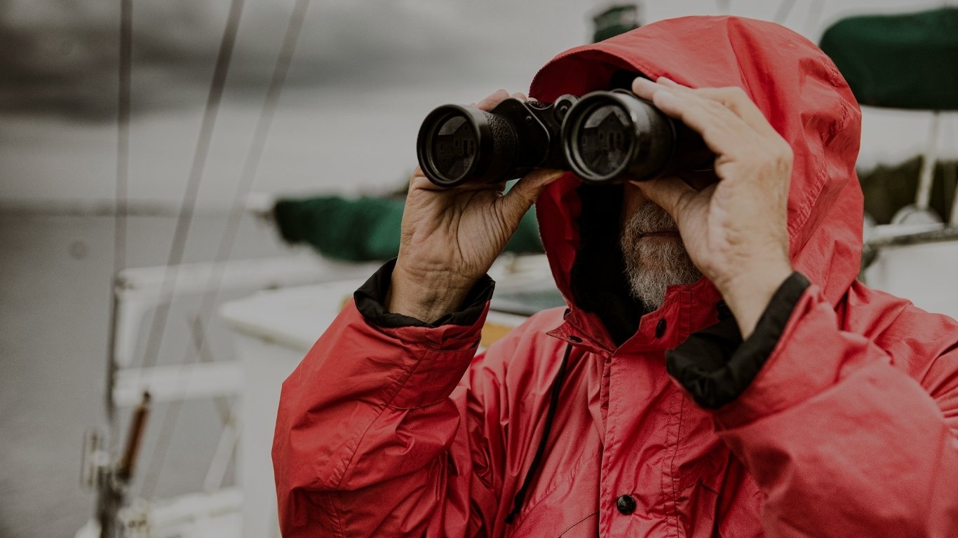 An elderly person wearing a red hooded jacket looking through binoculars, likely outdoors in a cold environment.