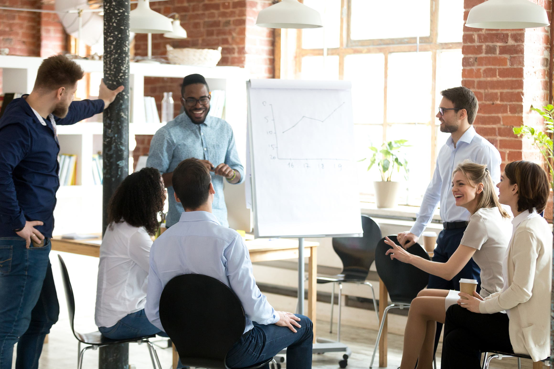 A group of diverse people in a business meeting or workshop in a bright, modern office space with brick walls. Some are sitting while others stand, engaging with a presentation on a whiteboard with a line graph. They are smiling and interacting.