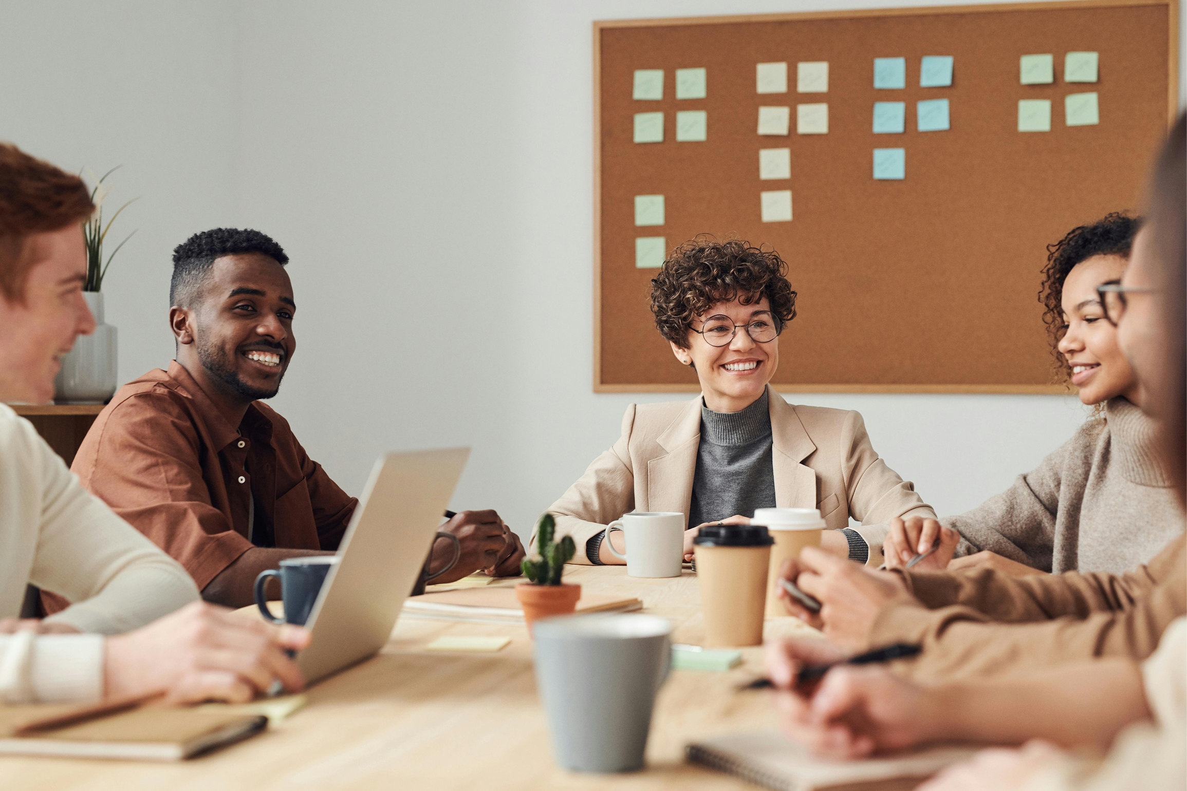 A diverse group of five people sitting around a table engaged in a meeting or discussion, with coffee cups, notebooks, and laptops, in a modern office with a corkboard filled with sticky notes on the wall.