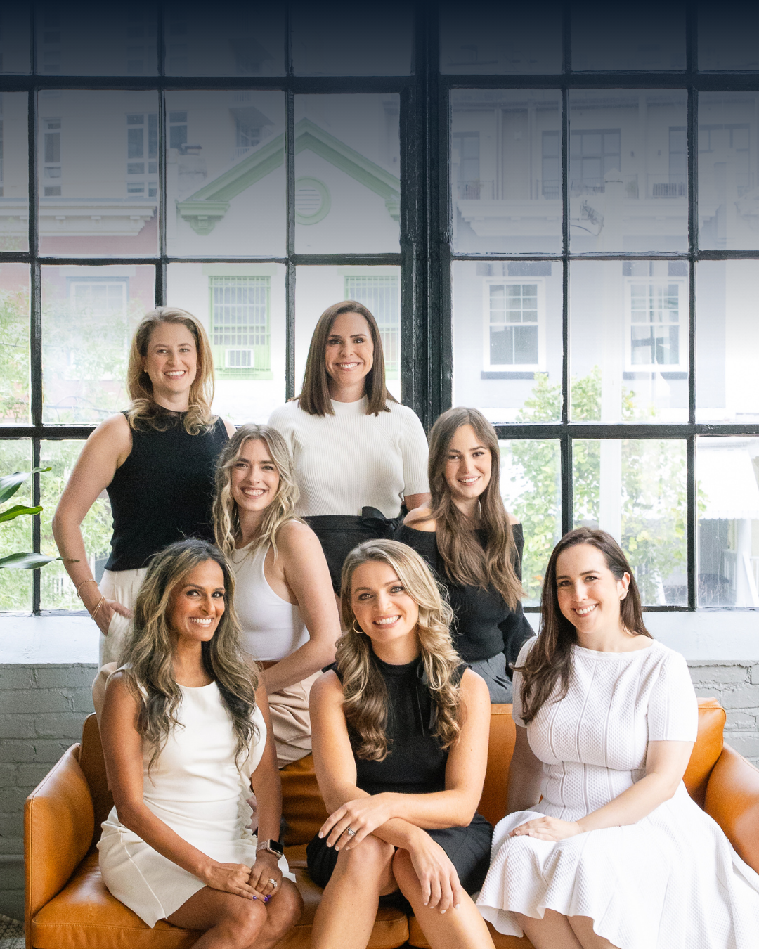 A group of eight women smiling and posing together in an indoor setting with large windows behind them.